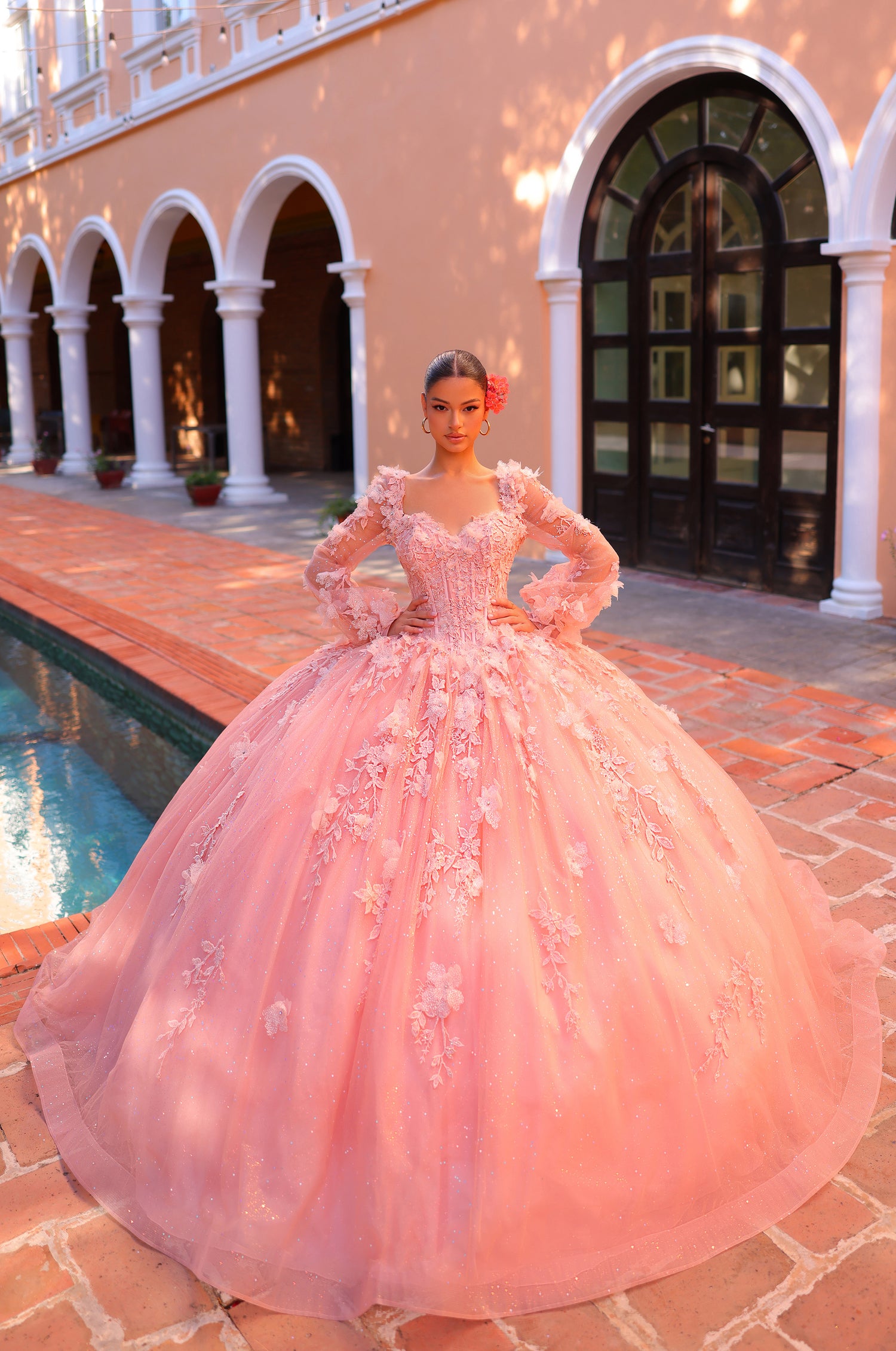 A blush pink, elaborate ball gown with 3D floral details sits on a model near arched columns and a pool.