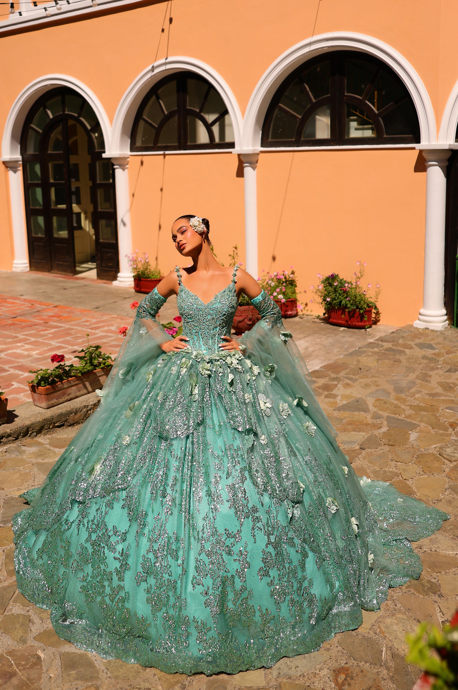 A dramatic sage green quinceañera ball gown with sparkling embellishments, worn on a stone courtyard with arched windows.