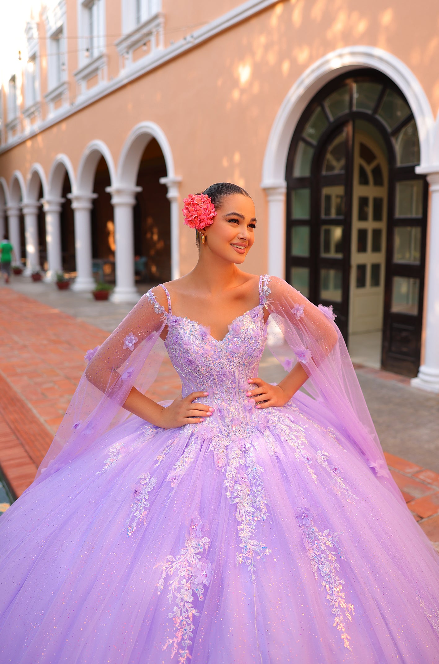 A woman in a lavender ball gown with floral accents poses in front of an arched building with white columns.