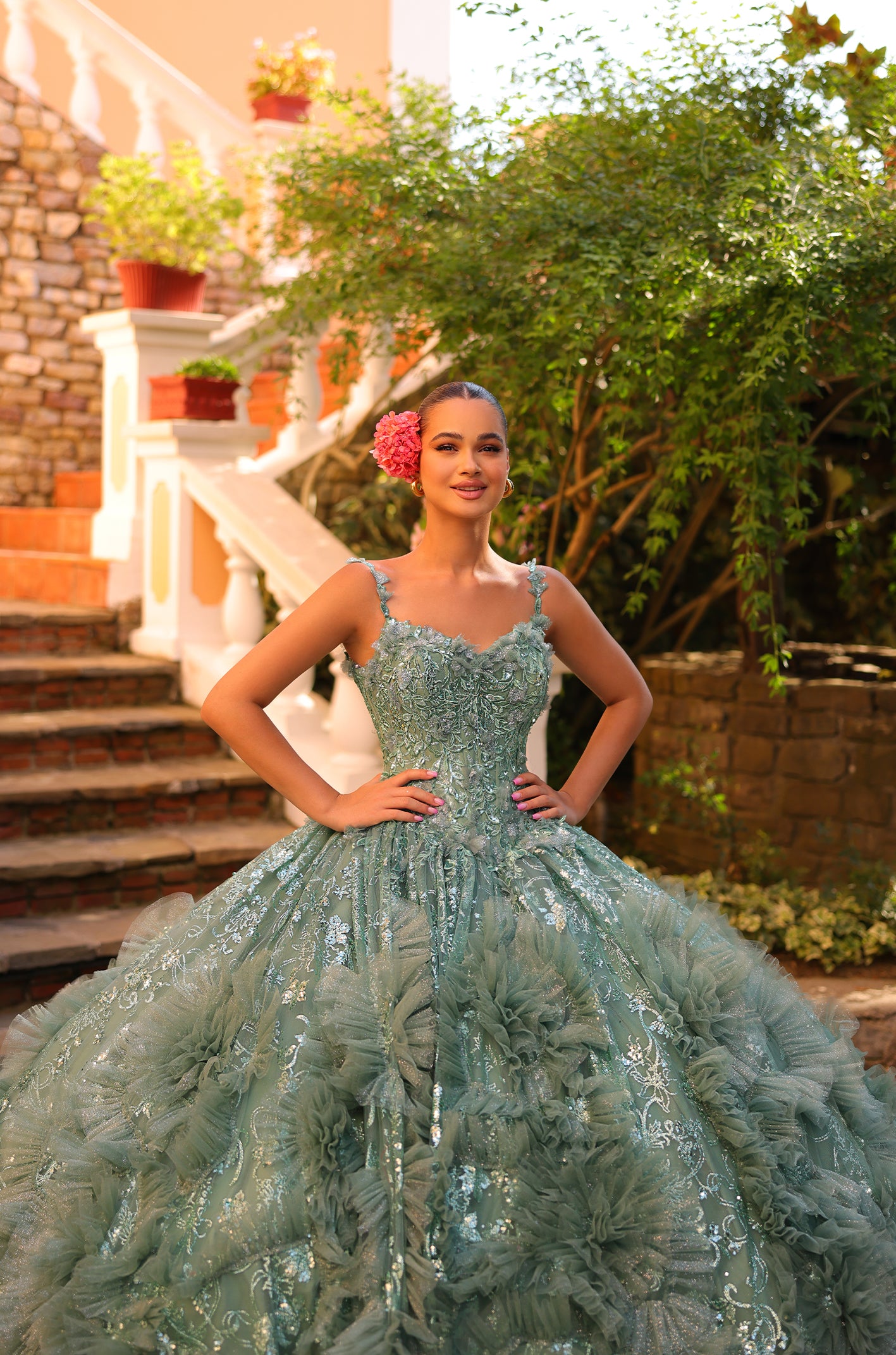 A young woman in a sage green, intricately embellished ball gown poses on stone steps with flowering plants.