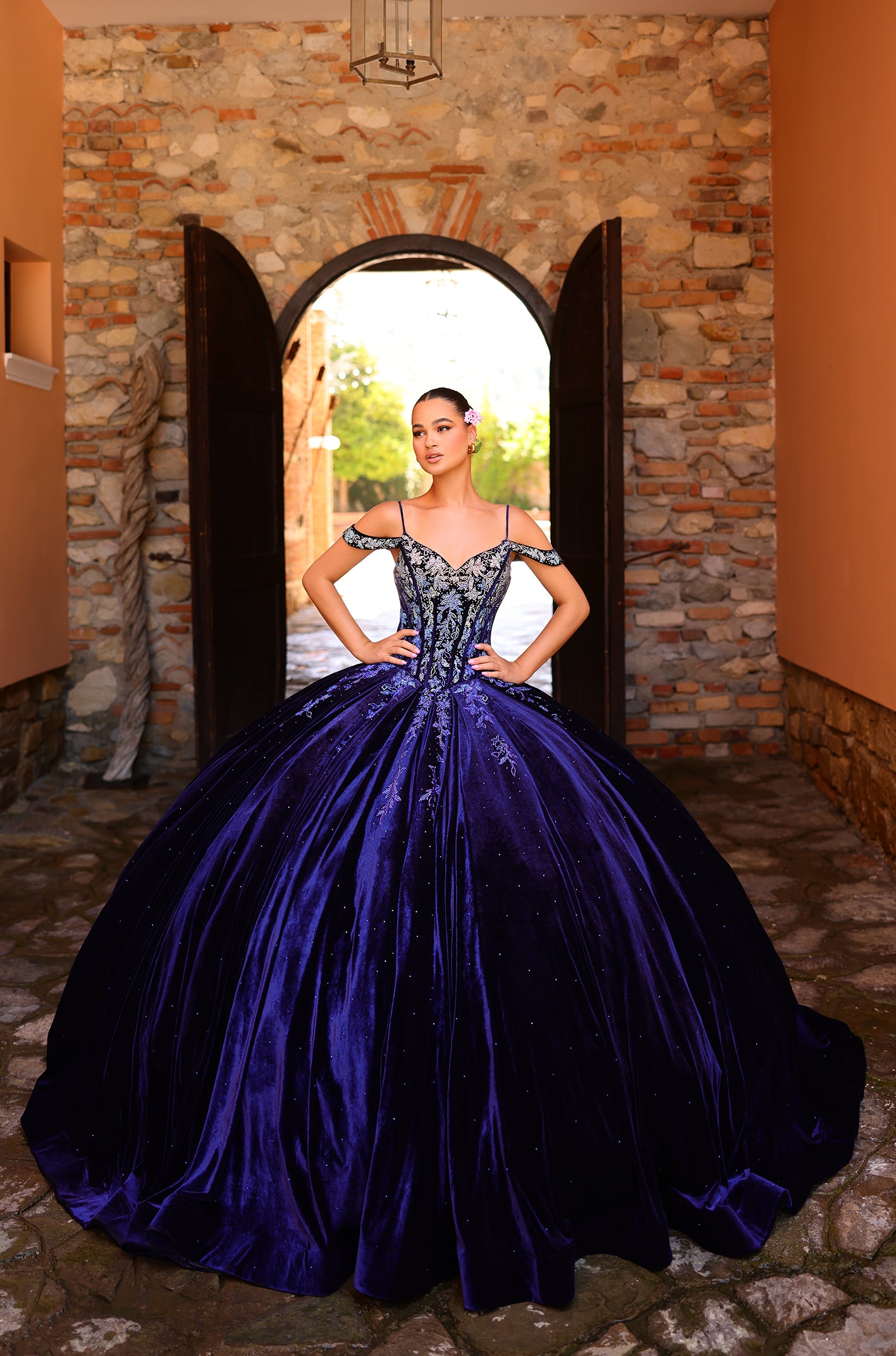 A woman in a royal blue ballgown poses confidently in a stone archway with her hands on her hips.