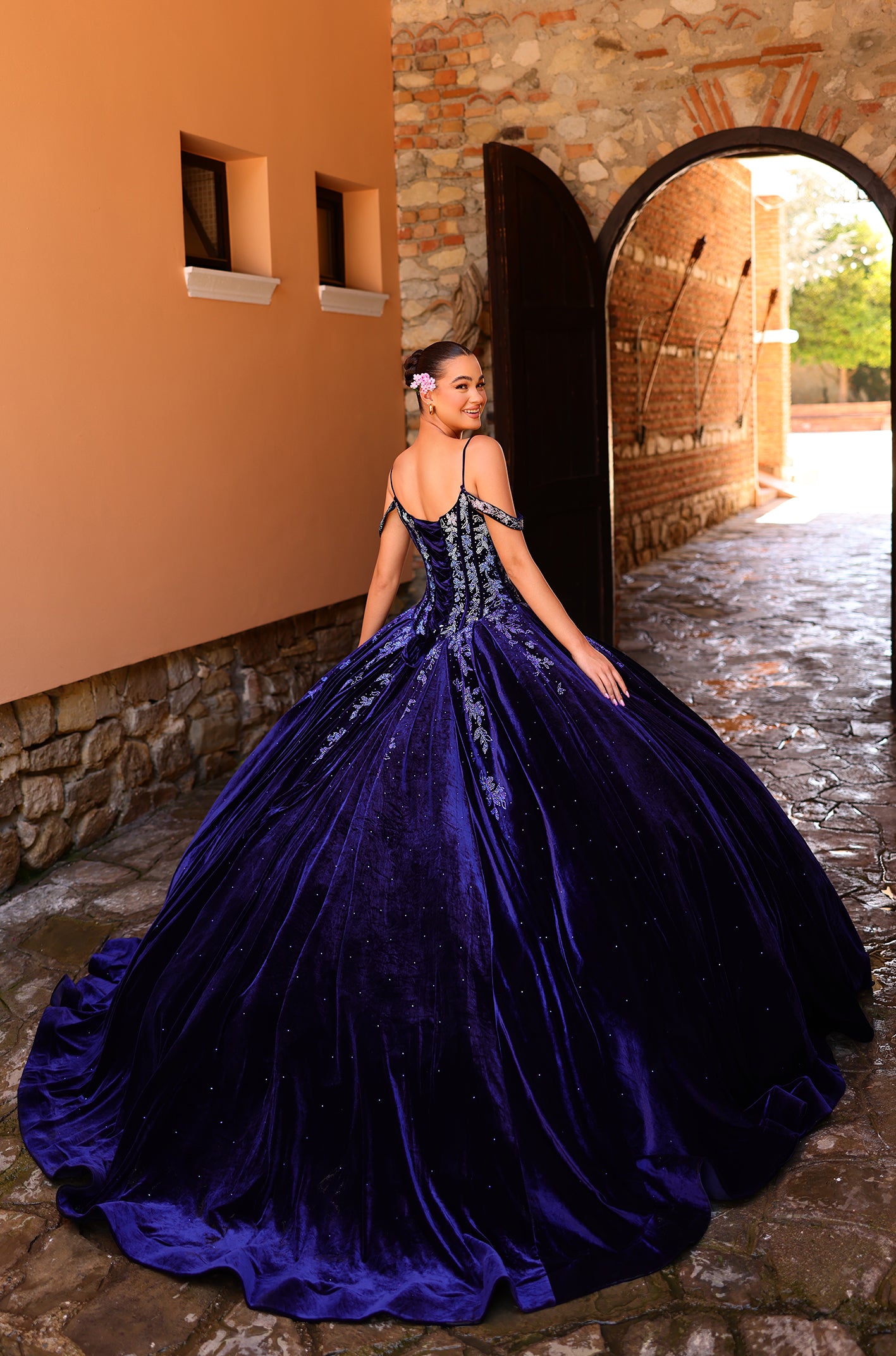 A woman in a royal blue ball gown with floral details poses in an arched stone corridor.