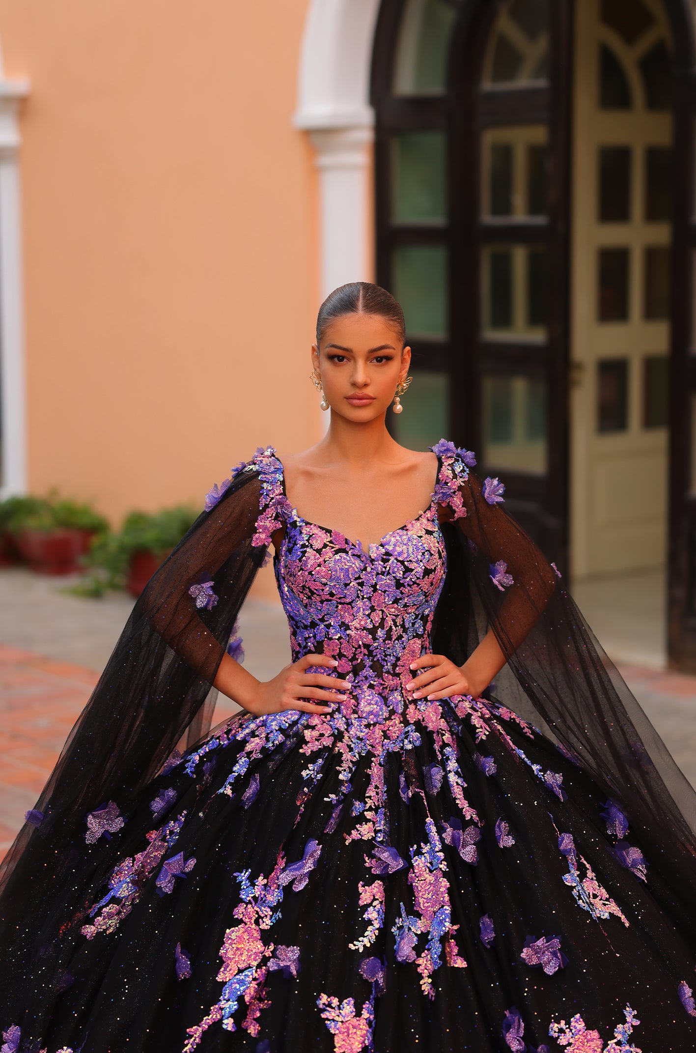 A model wears a black ball gown with purple and pink floral embroidery and sheer cape-like sleeves.