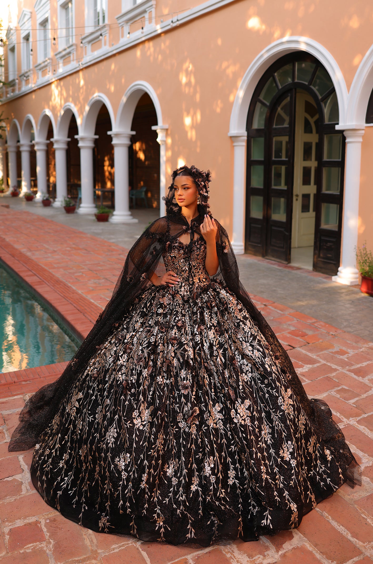 A woman wears an ornate black floral embroidered ball gown with a cape, standing by arched columns and a pool.