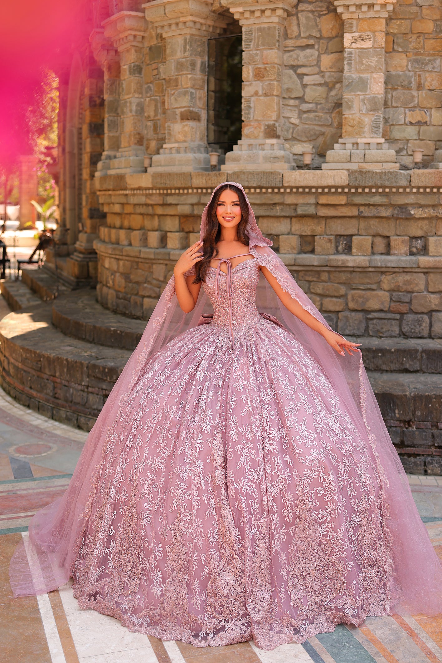 A woman in a lavender ballgown with a delicate lace cape stands before an ornate stone architectural backdrop.