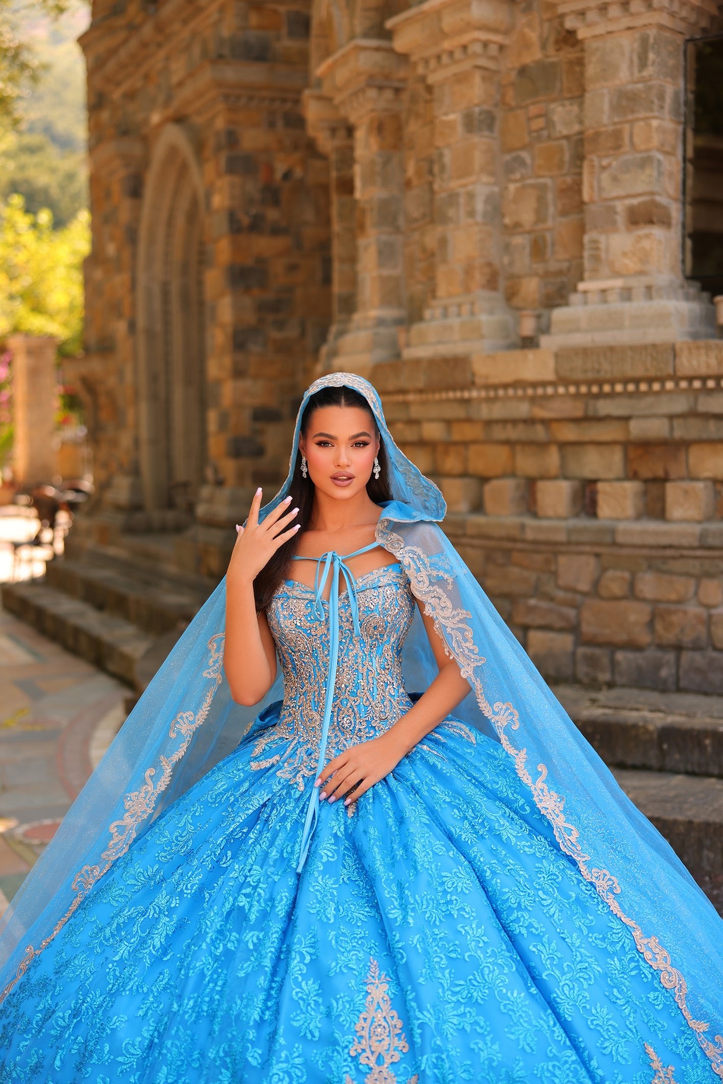 A woman in an ornate blue quinceañera gown with a matching veil poses before a stone architectural backdrop.