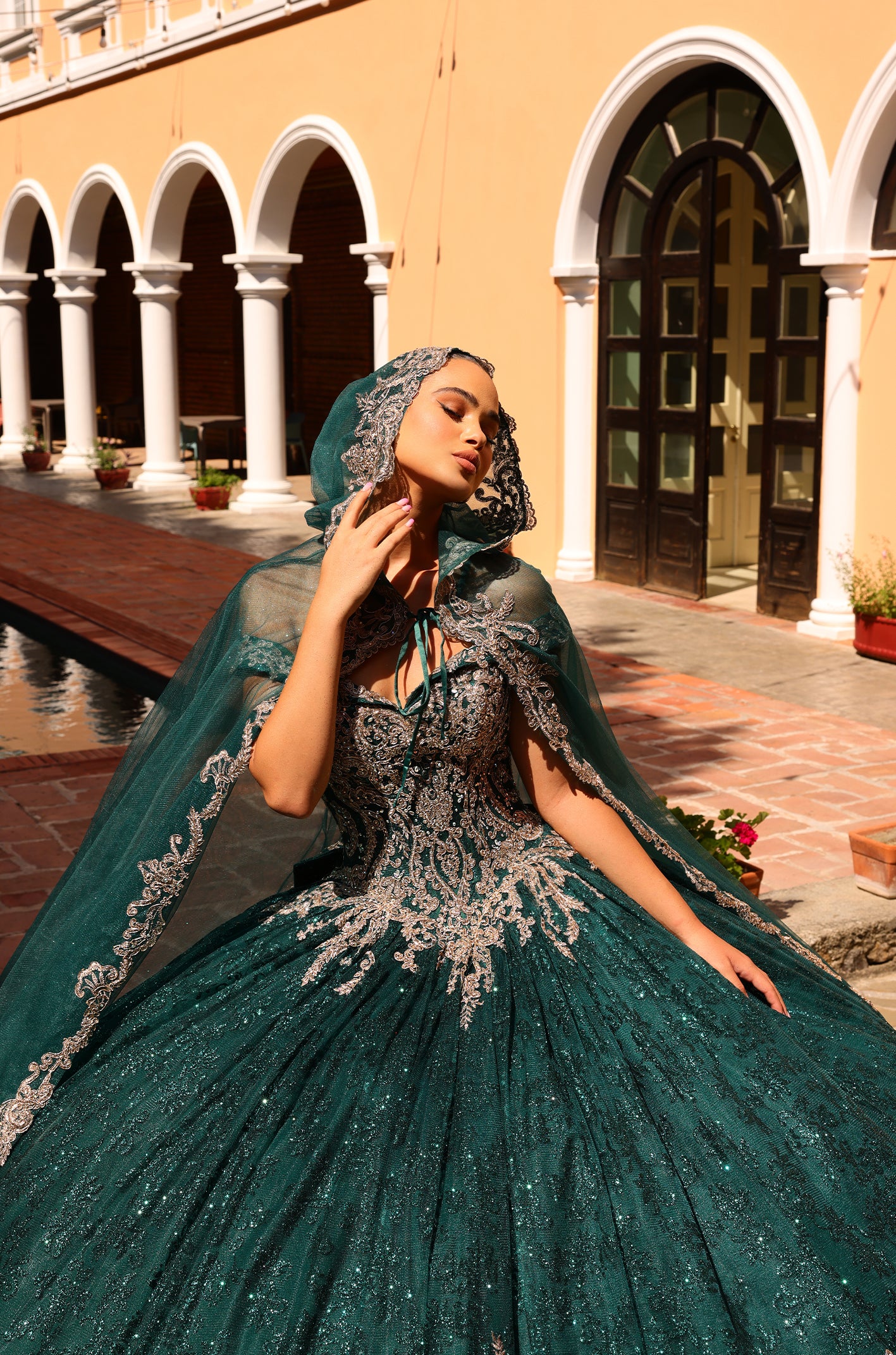 A model wears a sparkling emerald green ballgown with silver embroidery against an arched colonnade background.