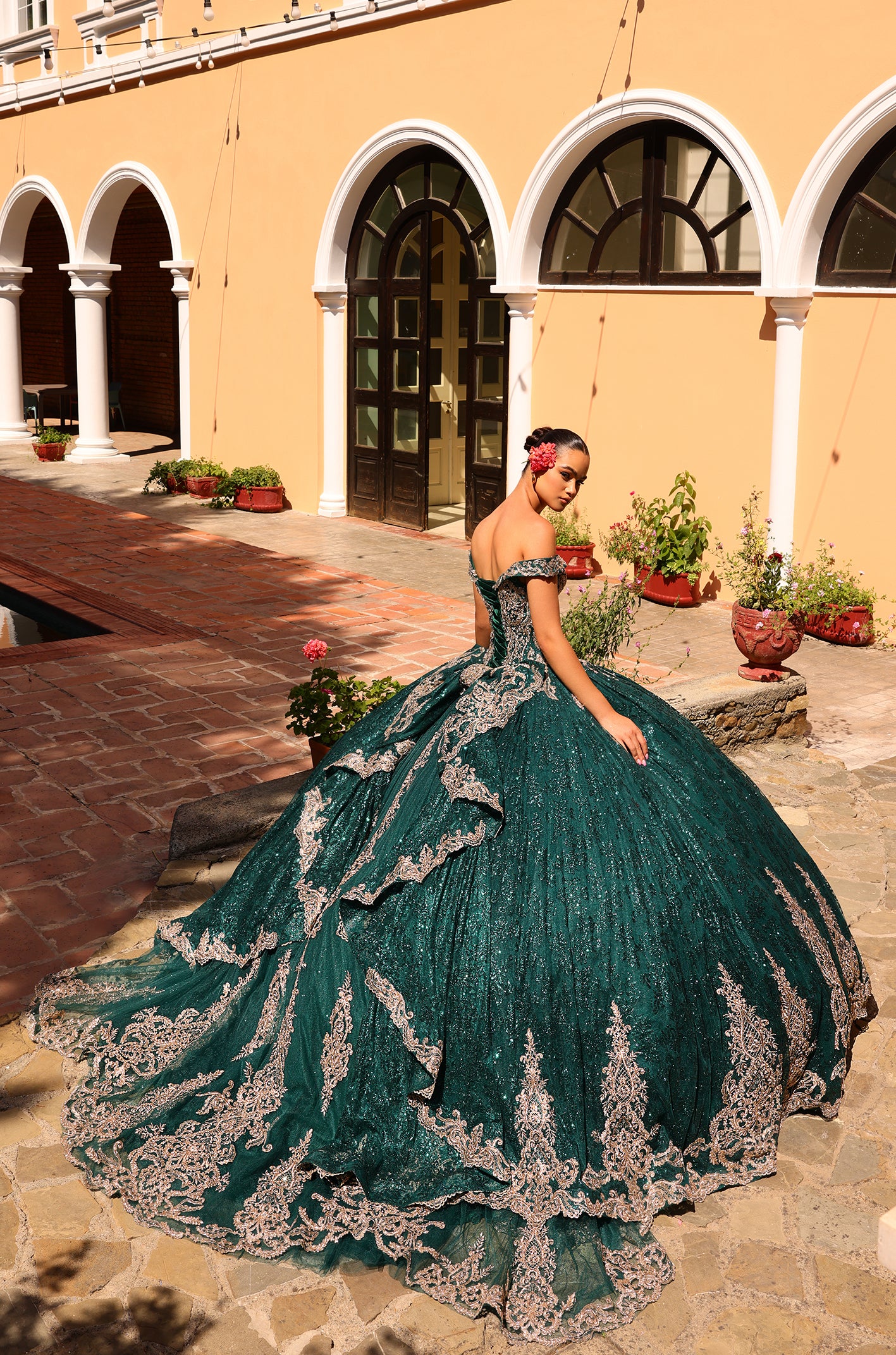 A woman wears an emerald green, off-shoulder ball gown with silver lace, posing in a sunlit courtyard with arched windows.