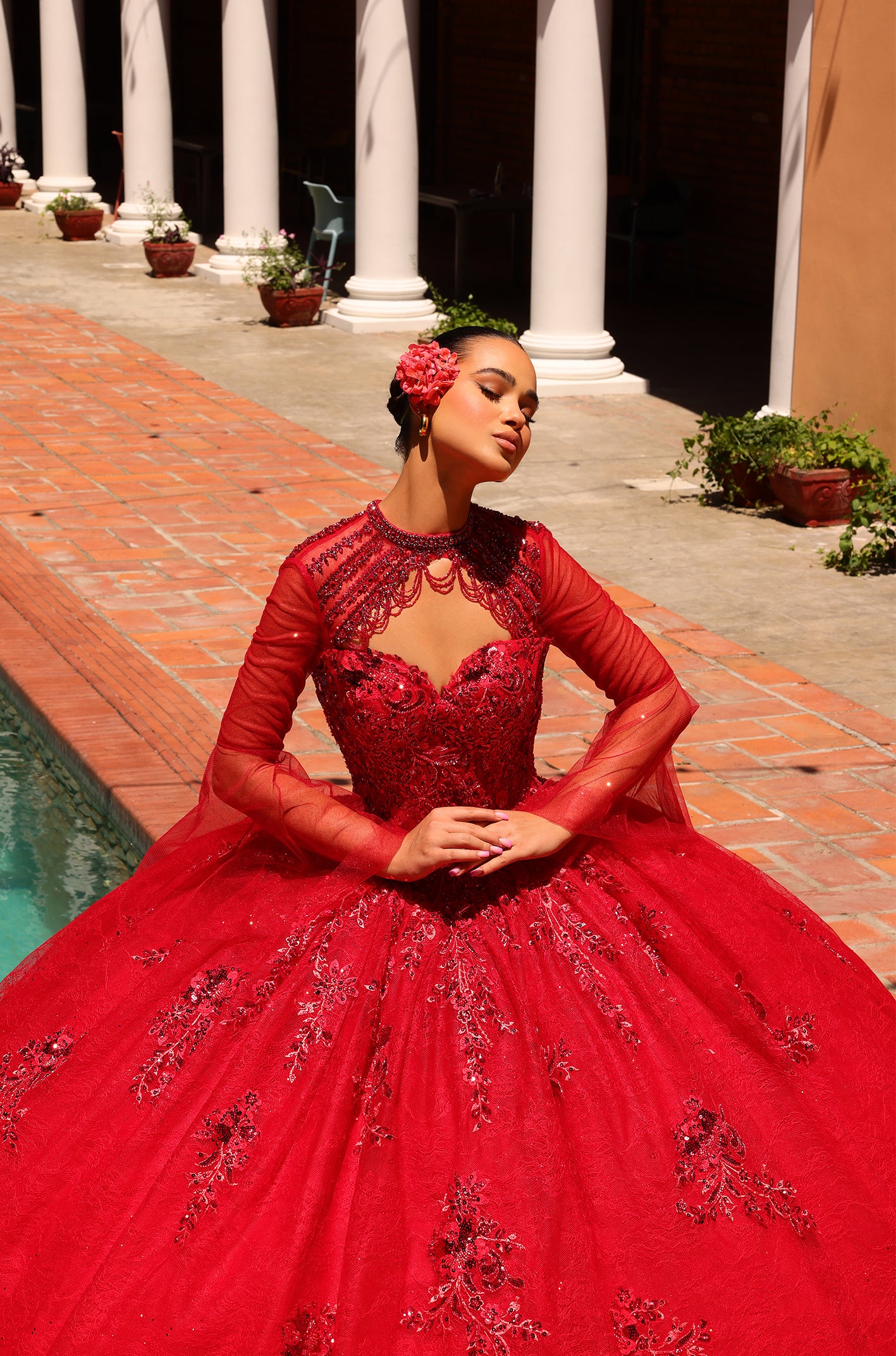 A woman in an ornate red ball gown poses elegantly beside columns and a pool, wearing a floral hair accessory.