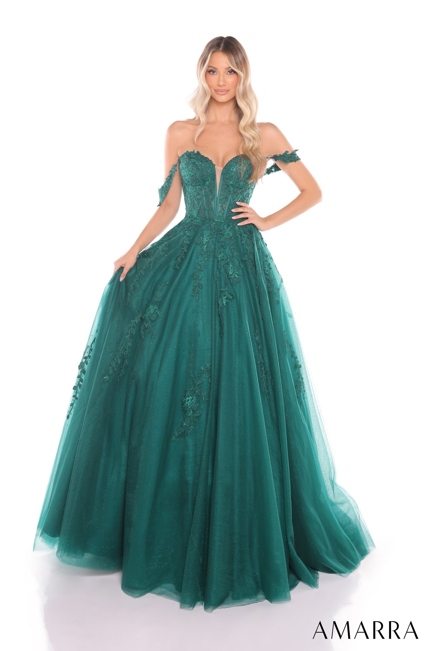 A blonde model wears an emerald ball gown with off-shoulder straps and intricate floral lace detailing.