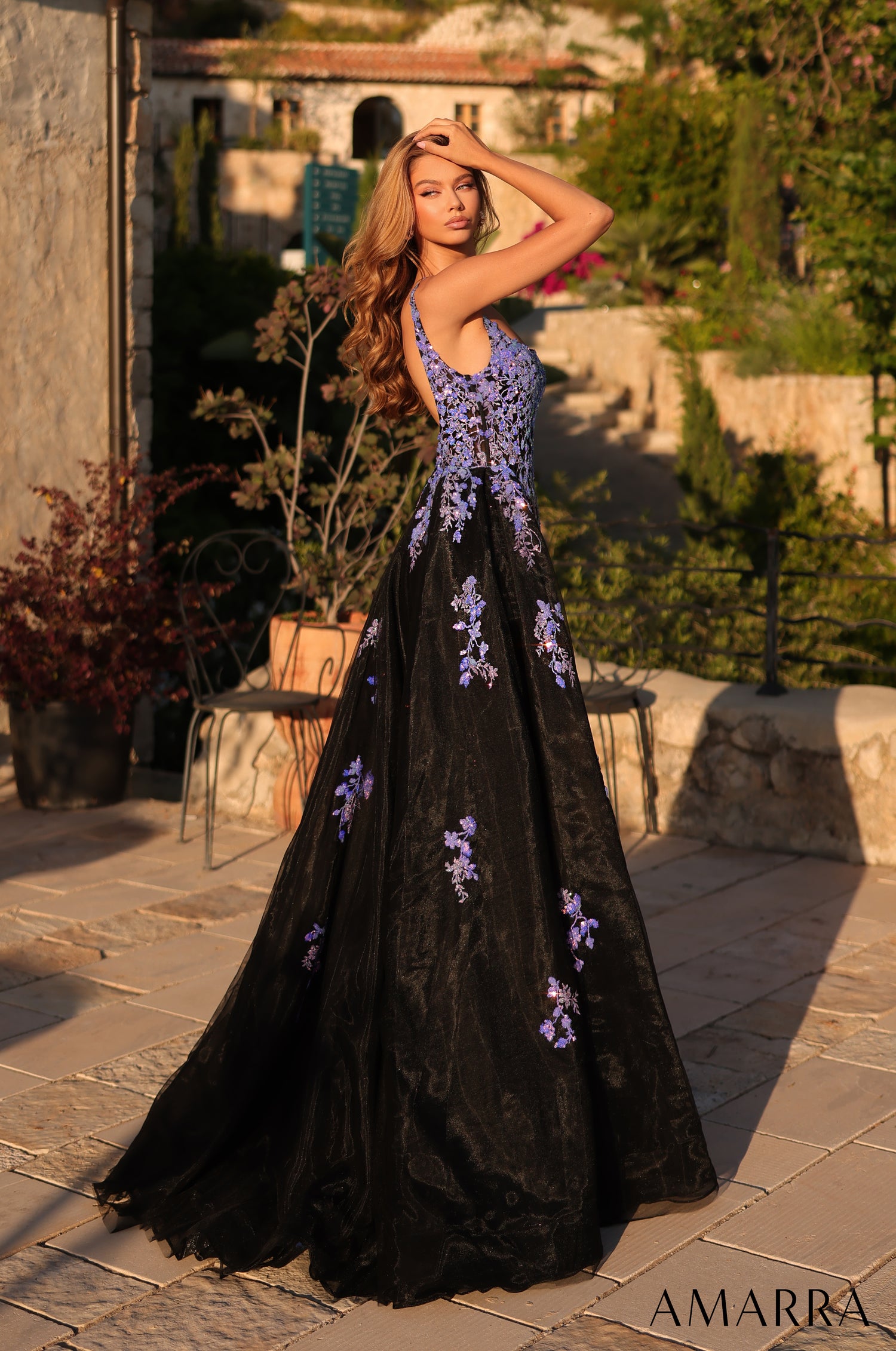 A model wears a black ball gown with purple floral embroidery in a Mediterranean-style garden setting.