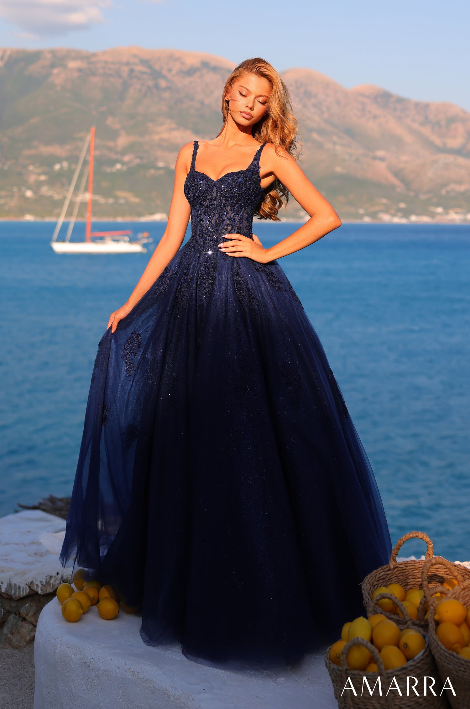 A model in a navy ball gown stands on a rocky shore with lemons, mountains, and a sailboat behind her.