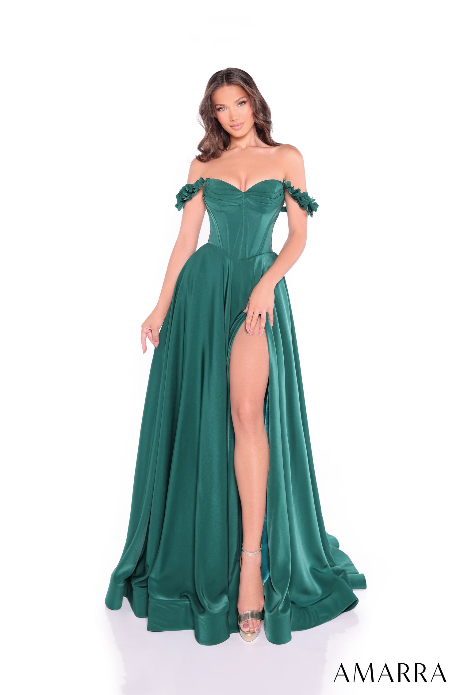A woman wears an emerald green off-shoulder gown with a high slit and flowing skirt on a white background.