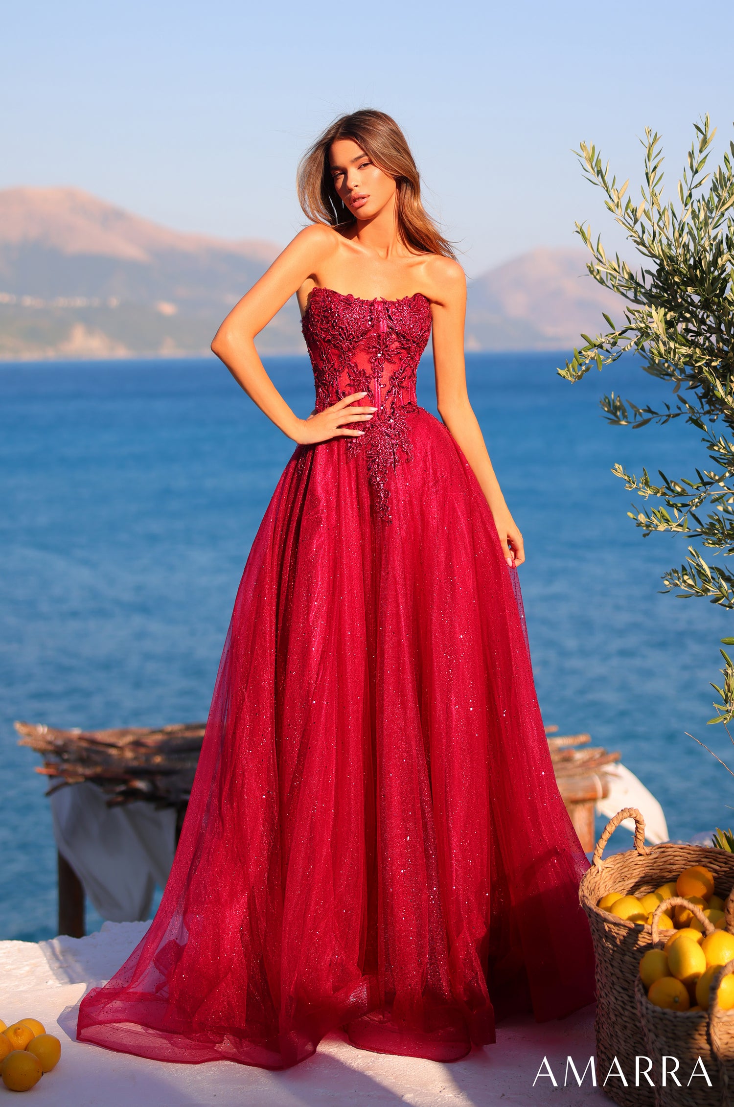 A woman wears a strapless wine-colored ball gown with sparkly embellishments against a seaside backdrop with lemons.