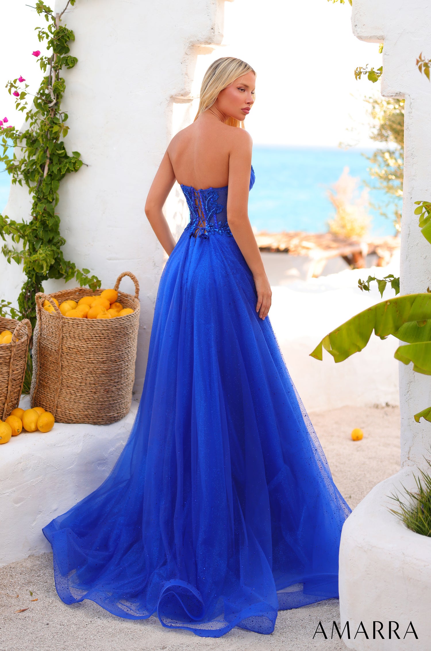 A woman wears a vibrant blue strapless ball gown with a corseted bodice against a Mediterranean coastal backdrop.