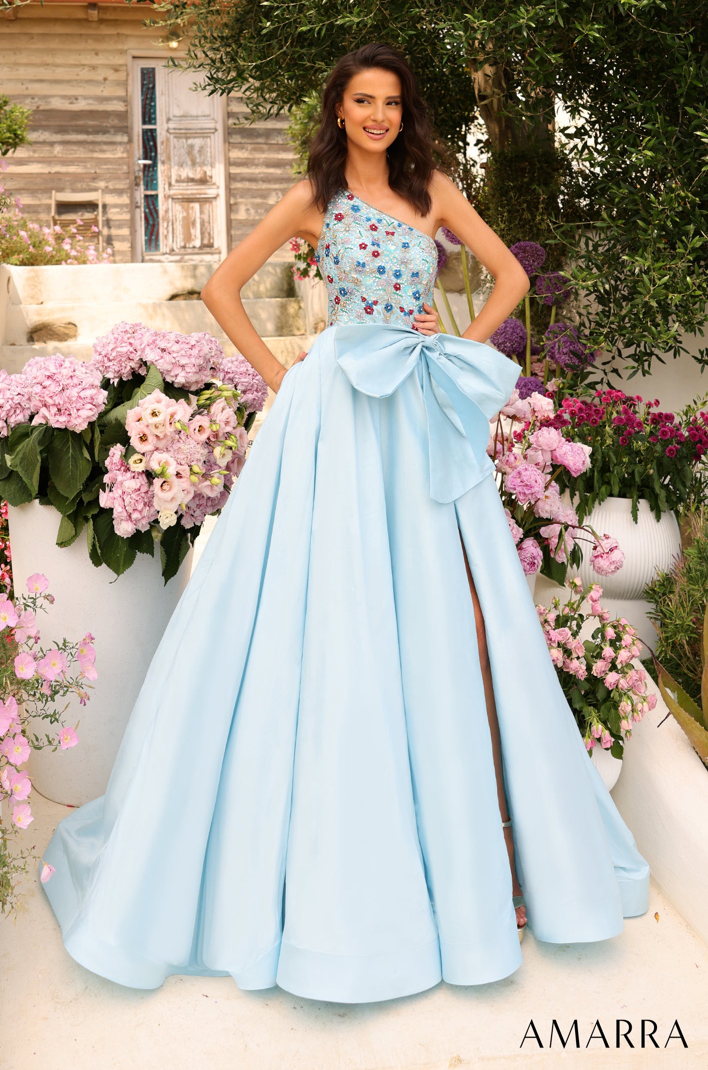 A woman wears a light blue one-shoulder ball gown with floral embellishments and a large bow, surrounded by pink flowers.