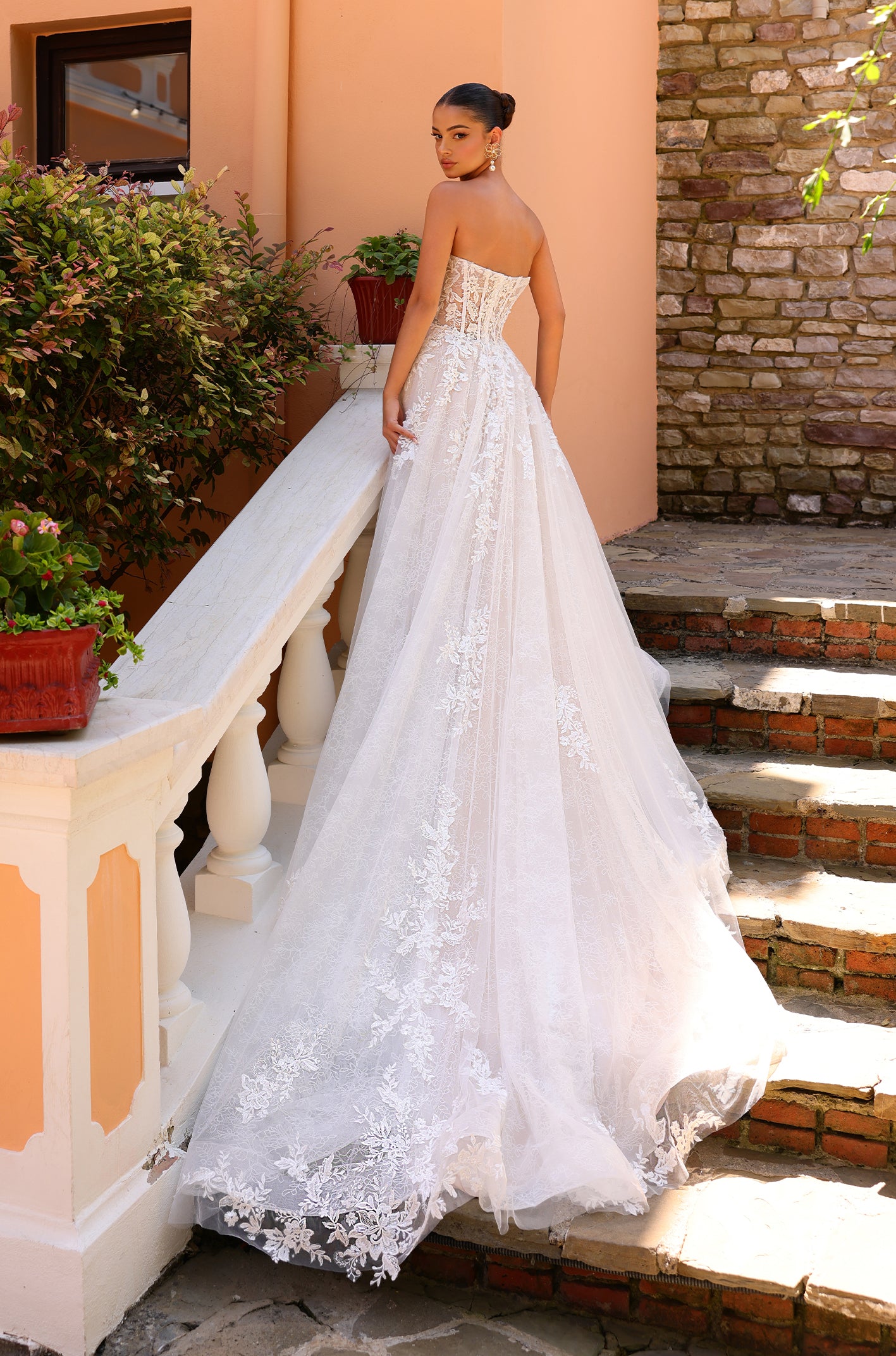 A bride in a white strapless lace ballgown stands on stone steps, her elaborate dress trailing behind her.
