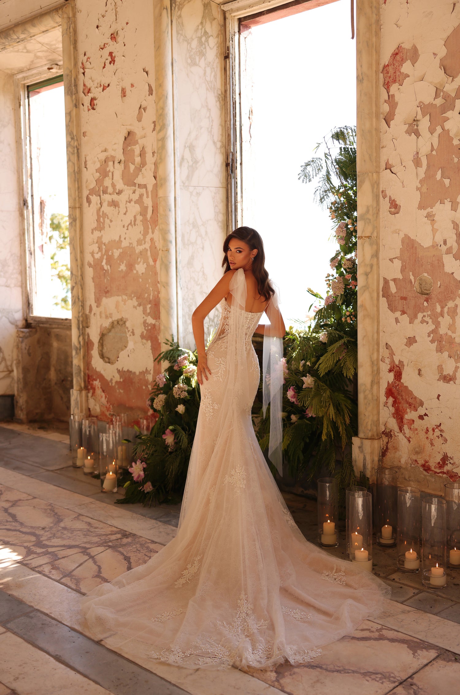 A bride in an elegant lace mermaid wedding gown stands in a distressed room with candles and floral decor.