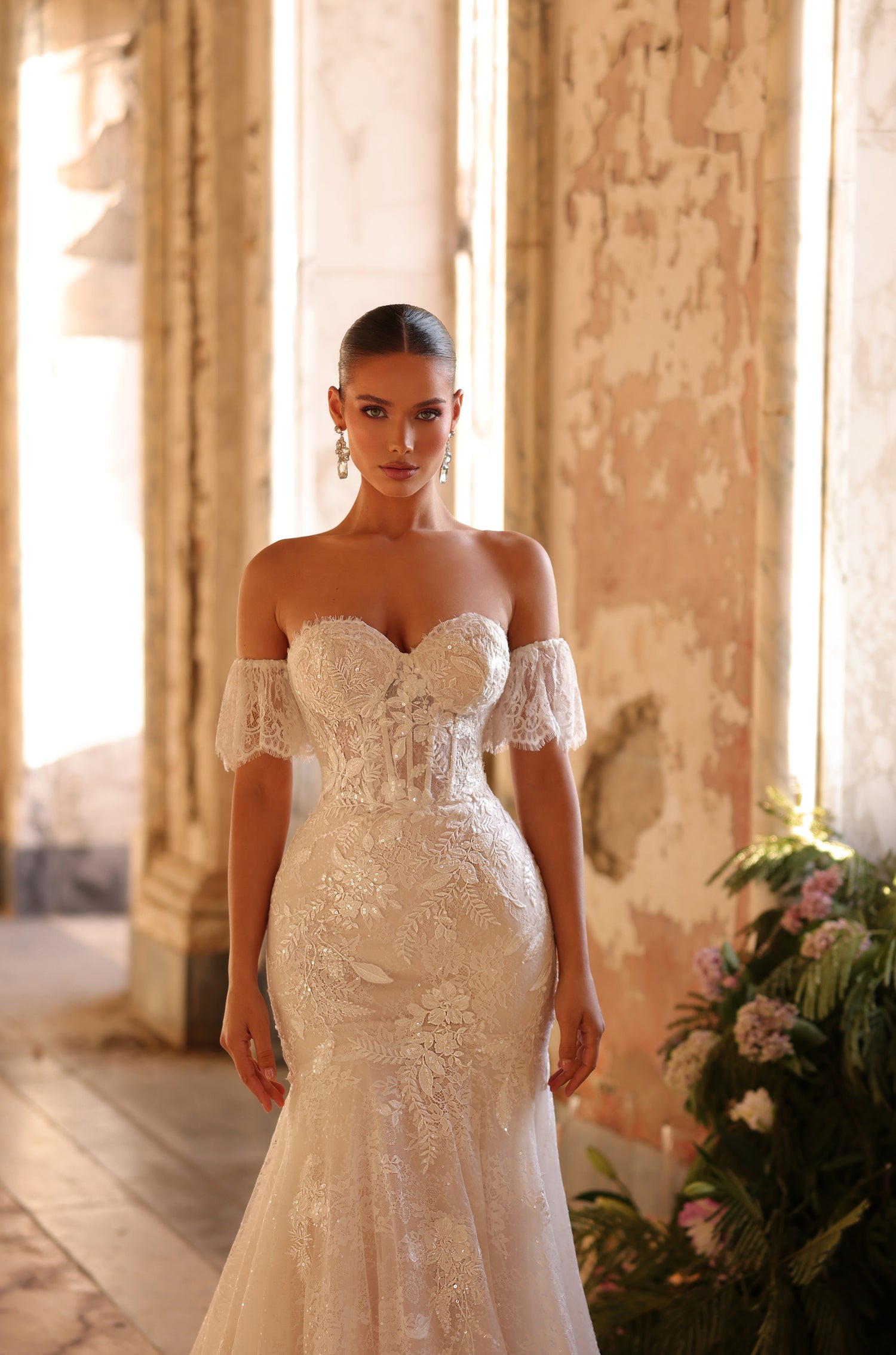 A bride in an off-shoulder lace mermaid wedding gown stands in a sunlit, vintage room with floral decor.