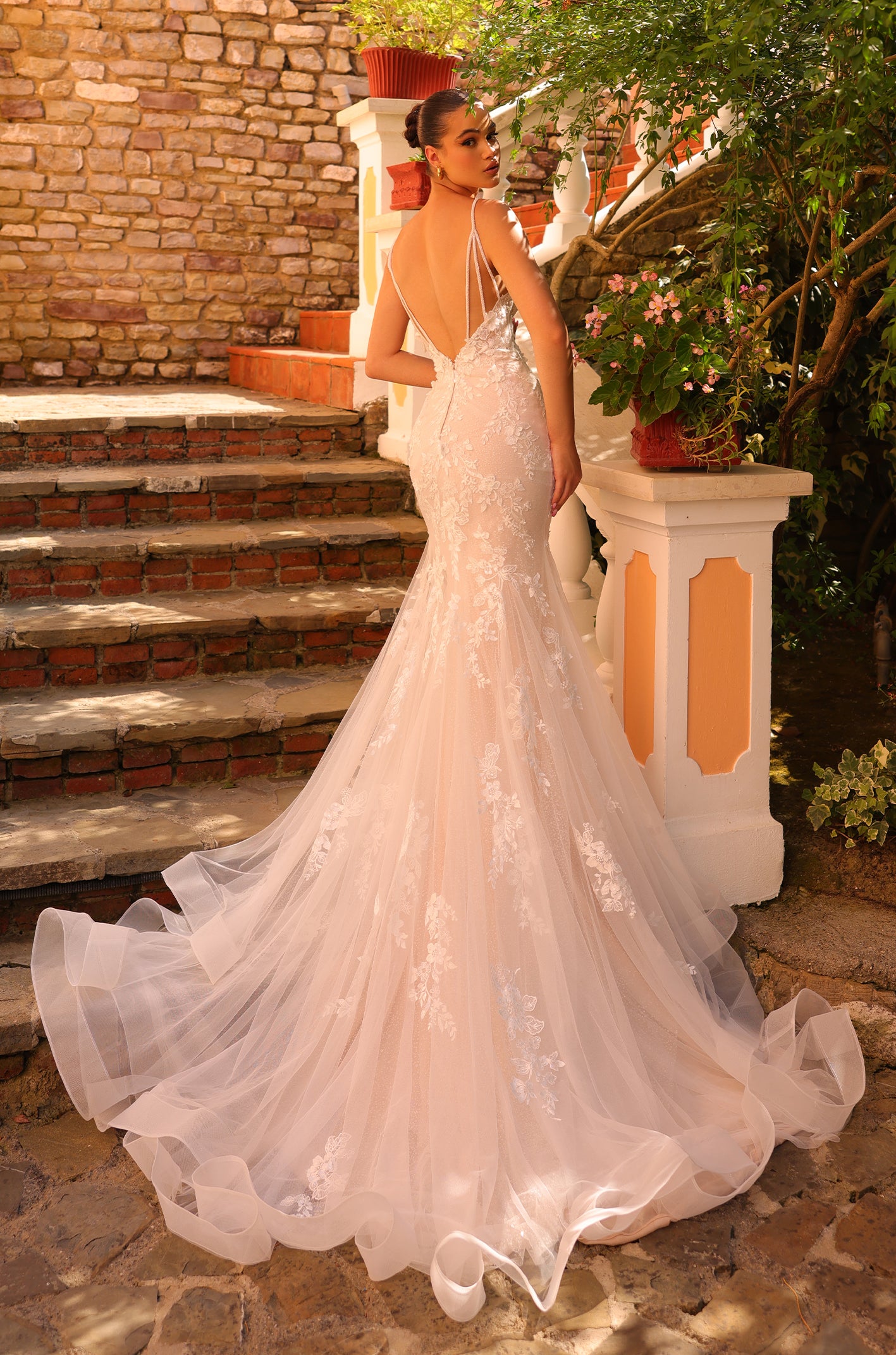 A bride in a blush lace mermaid wedding gown stands on brick steps with a flowing tulle train.