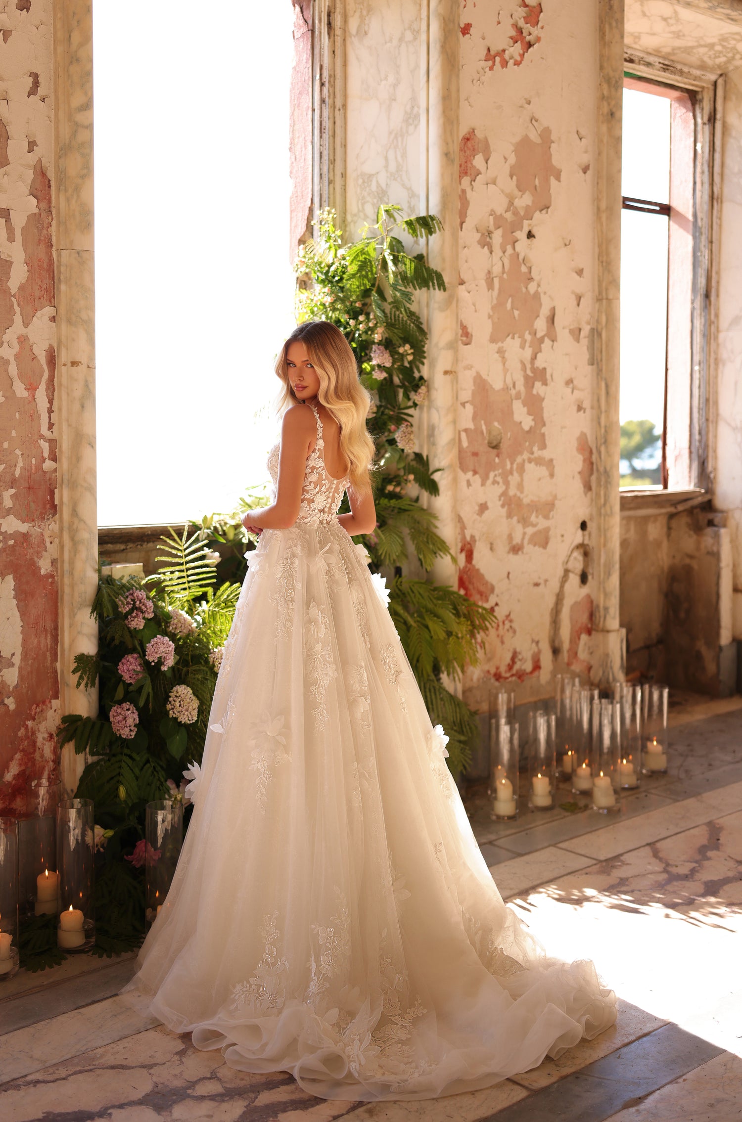 Bride in lace ballgown stands by window with candles and greenery in weathered, romantic interior.