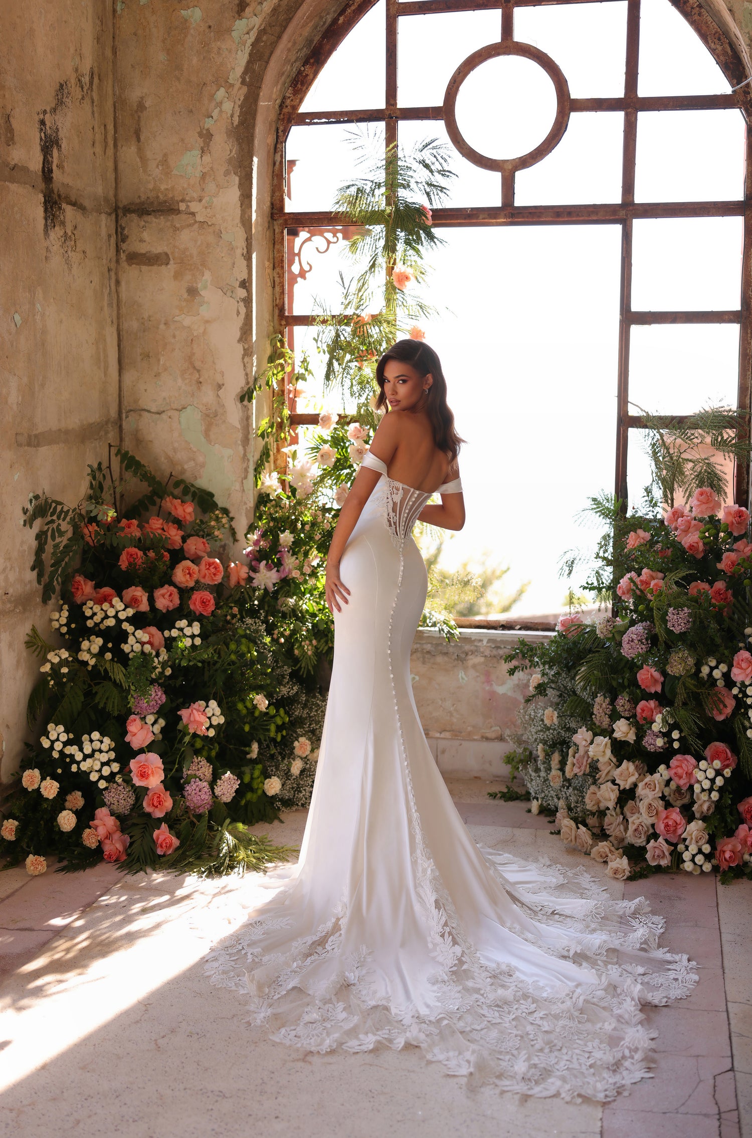 A bride in an off-shoulder white mermaid gown stands by an arched window surrounded by pink and white flowers.
