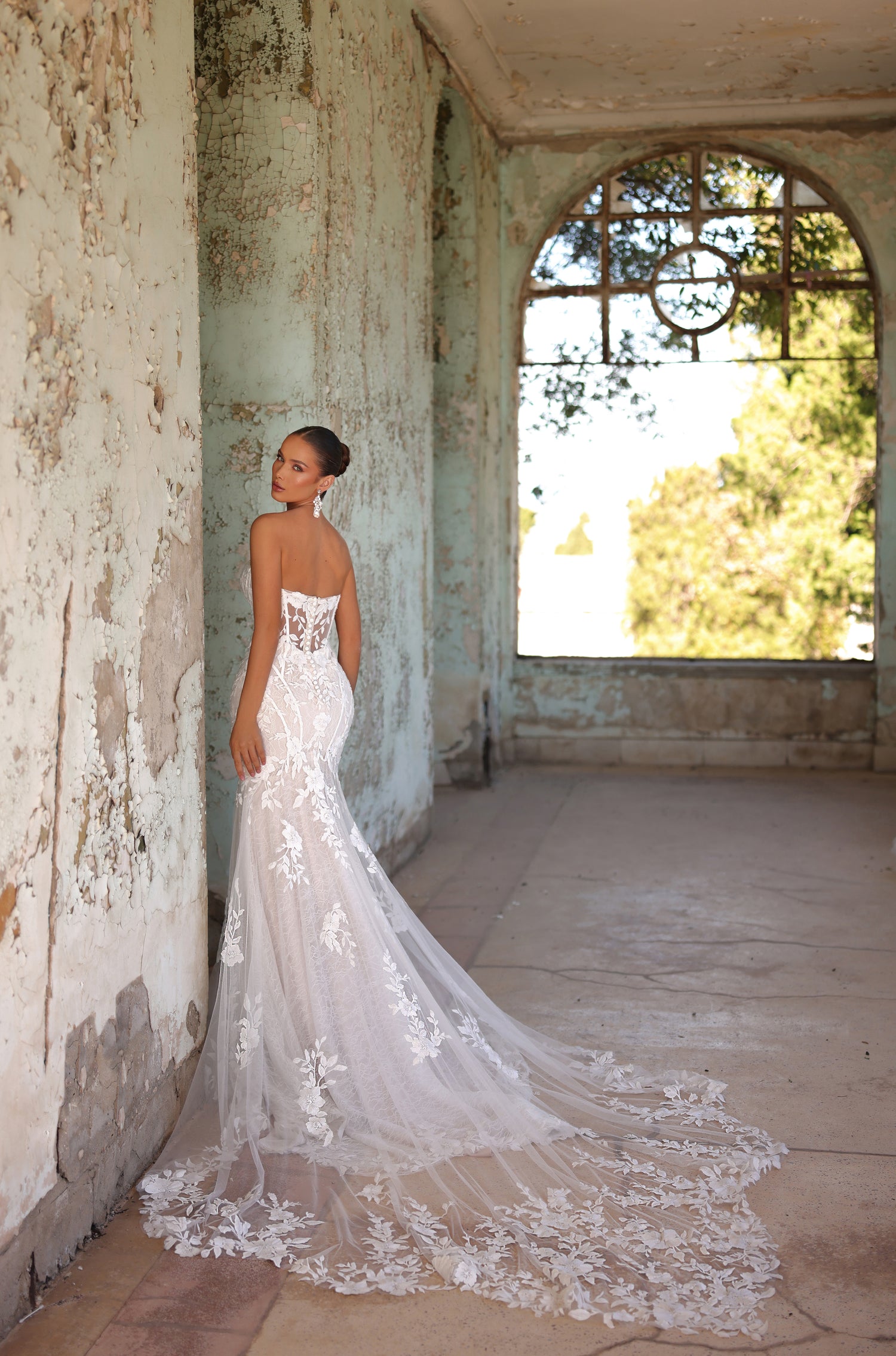 A bride in a lace mermaid wedding dress stands in a weathered room with an arched window.