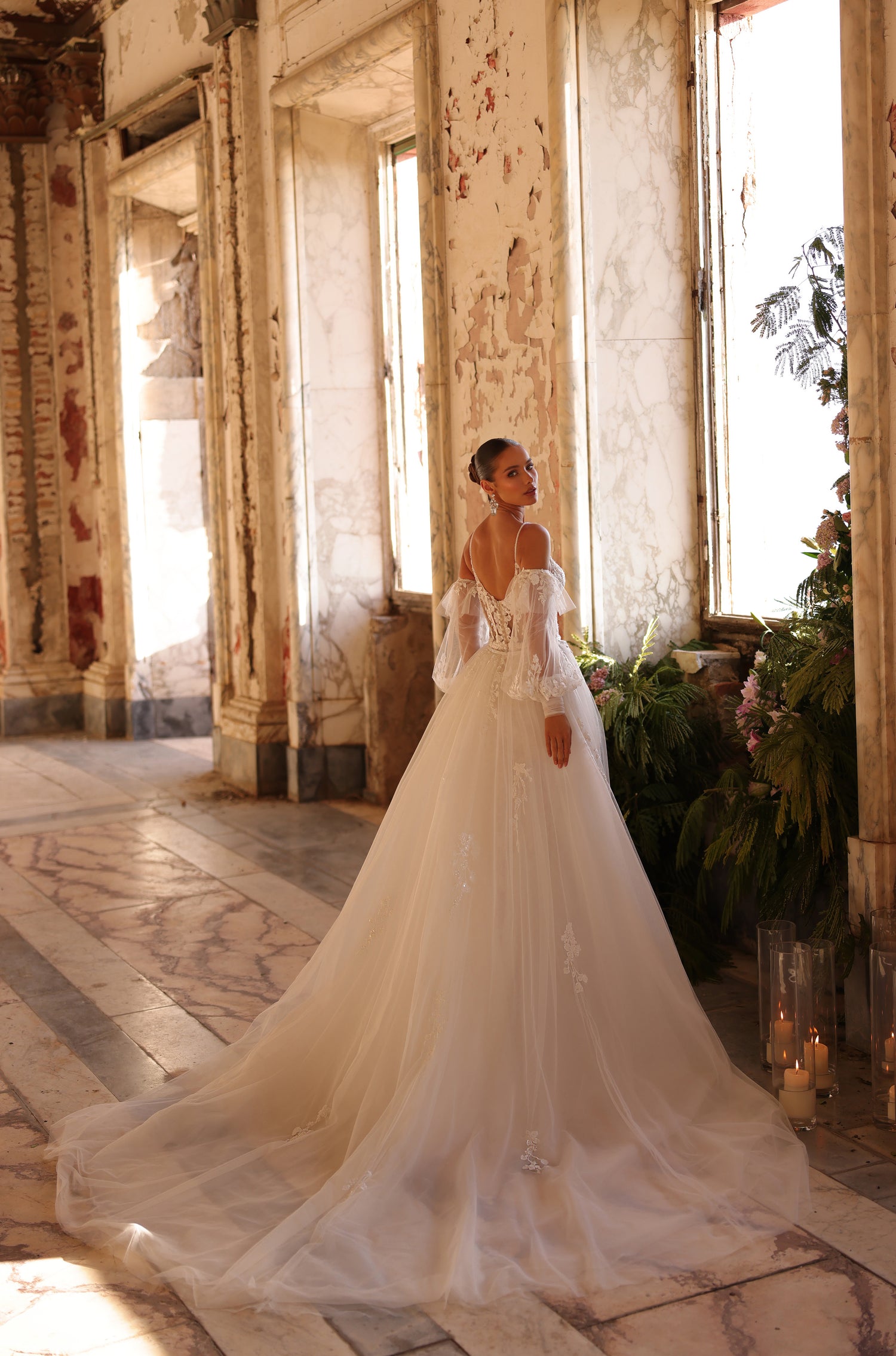 A bride in an off-shoulder ballgown stands in a sunlit, distressed room with candles and plants.