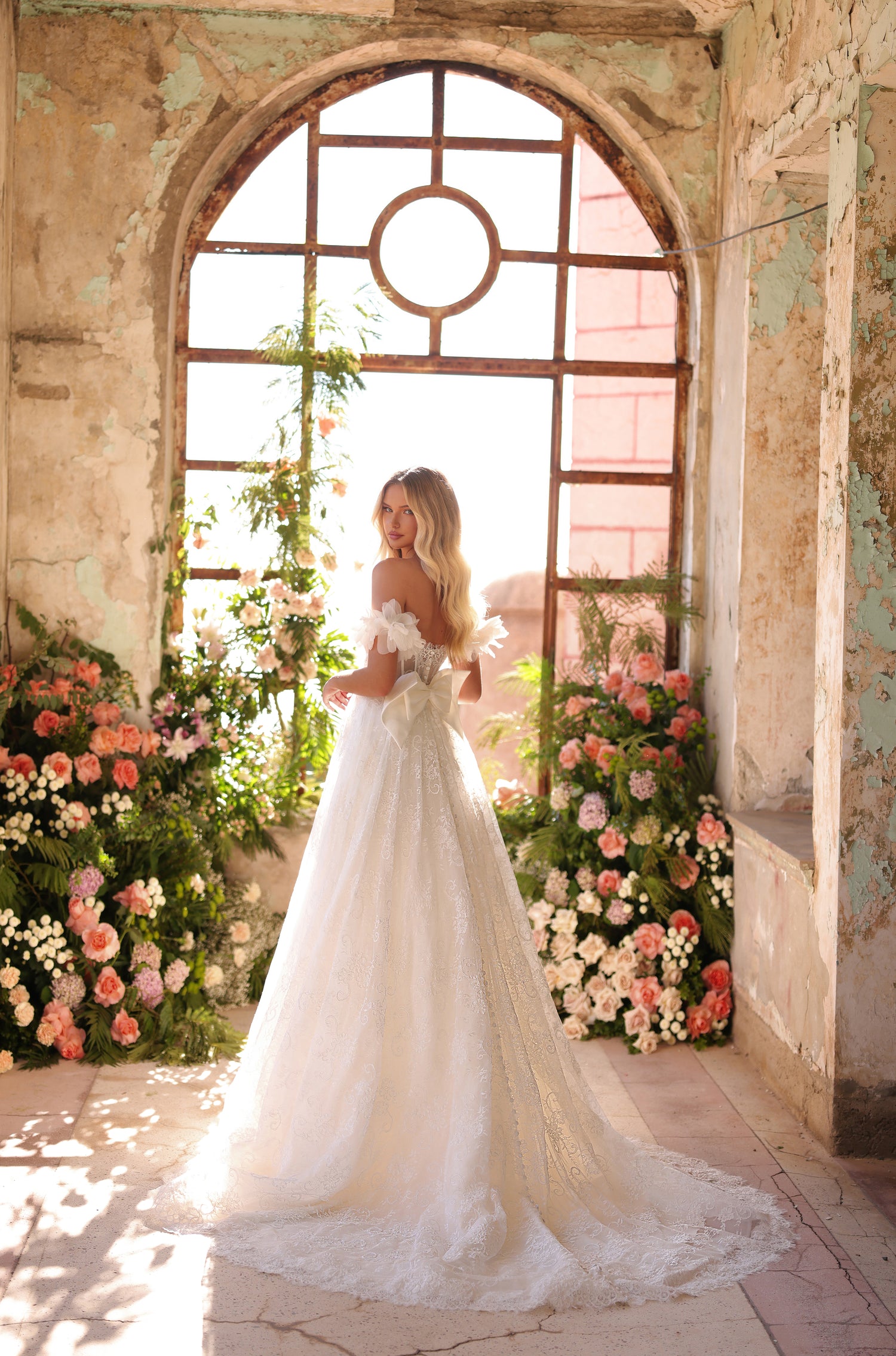 A bride in an off-shoulder white lace gown stands in a floral-filled, rustic arched window with soft light.