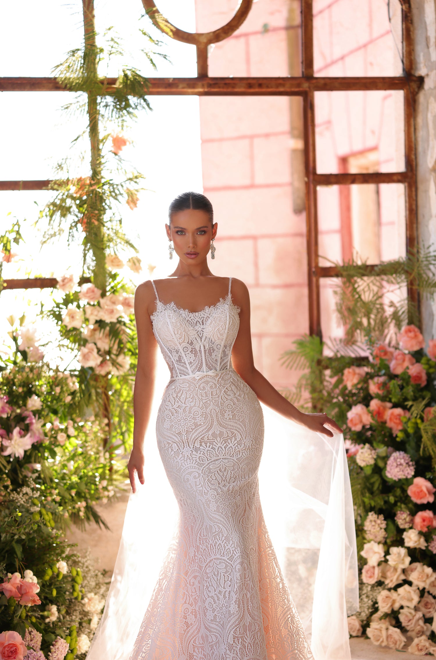 A bride in an intricate white lace mermaid wedding gown stands amid blush roses in a sunlit greenhouse.