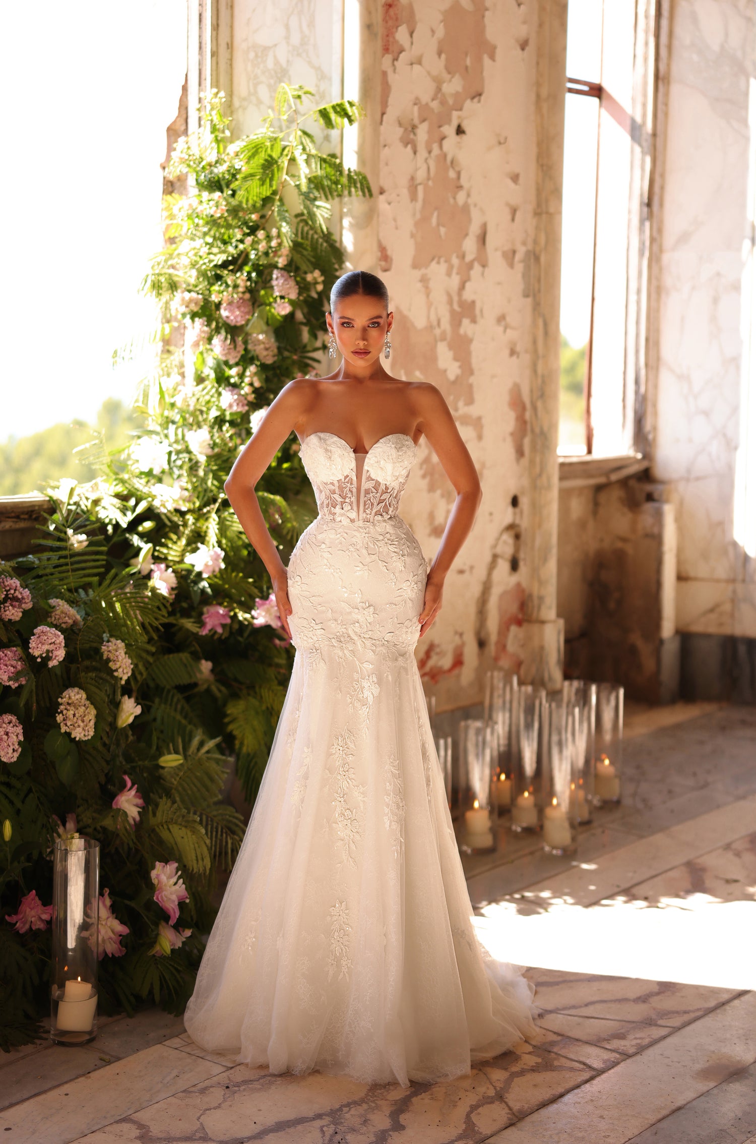 A bride in a strapless, mermaid-style white wedding gown stands in a rustic room with flowers and candles.