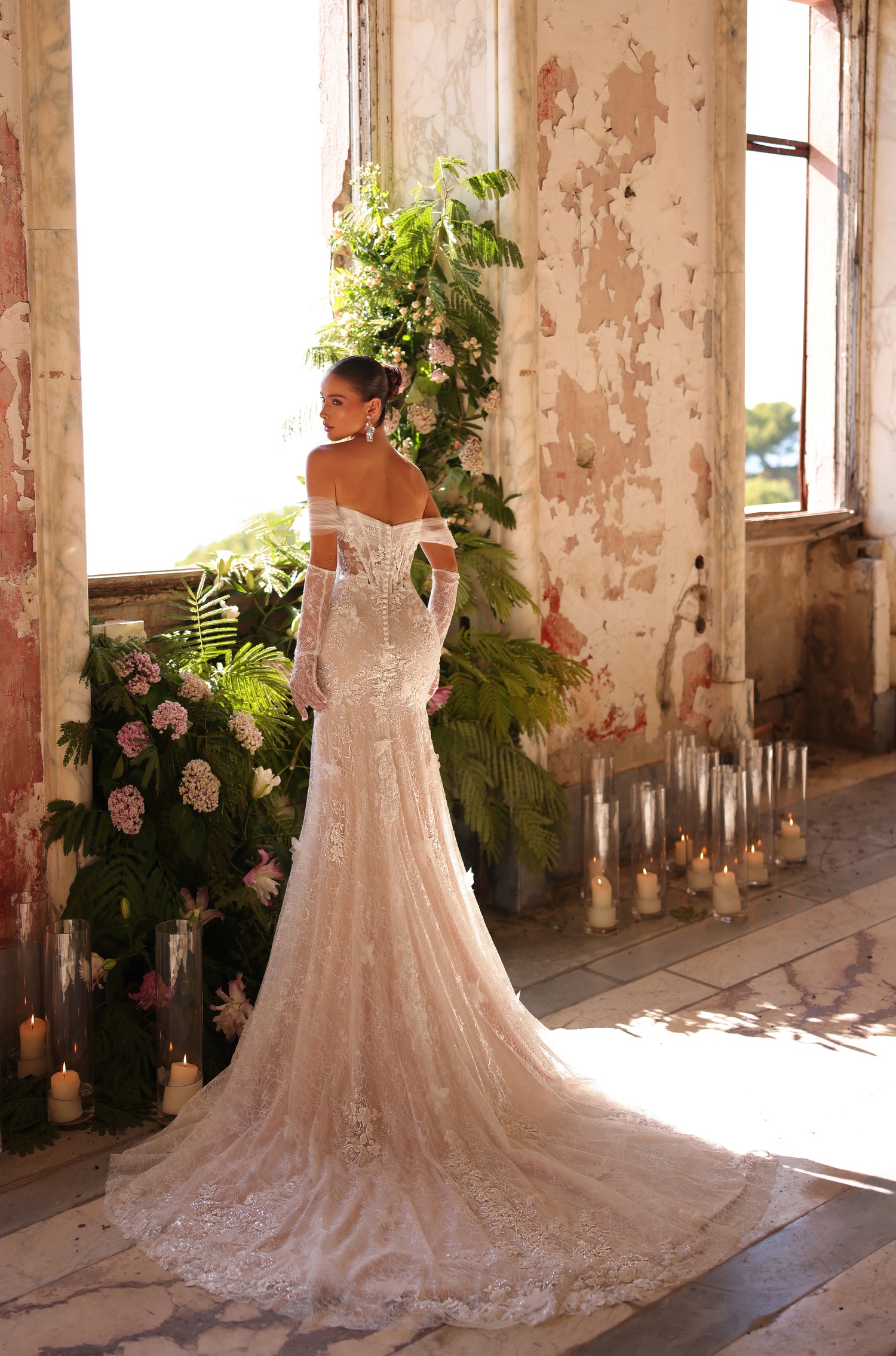 Bride in lace mermaid wedding gown stands in distressed room with candles and greenery.