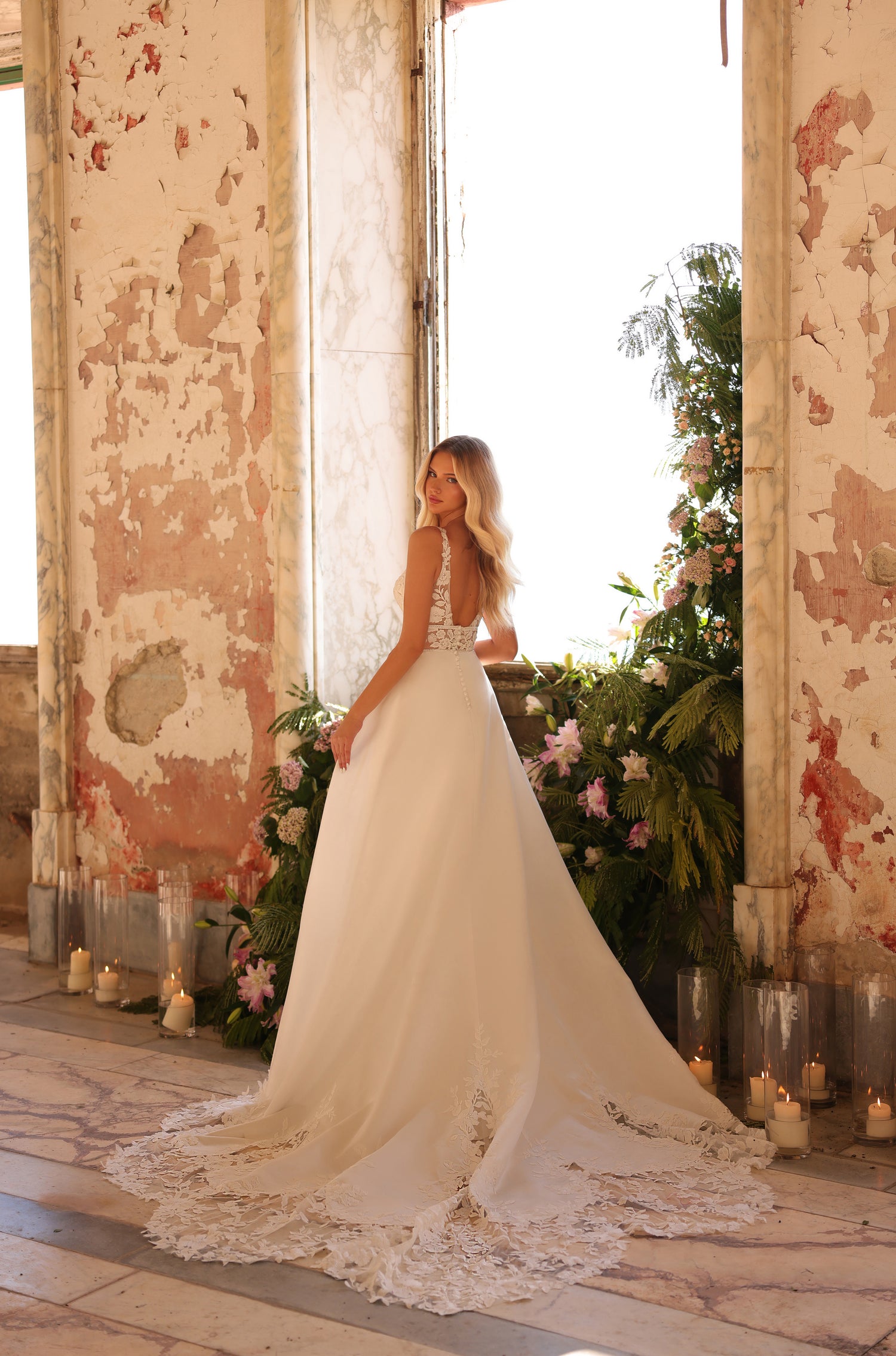 A bride in a lace wedding gown stands in a vintage room with candles and tropical flowers.