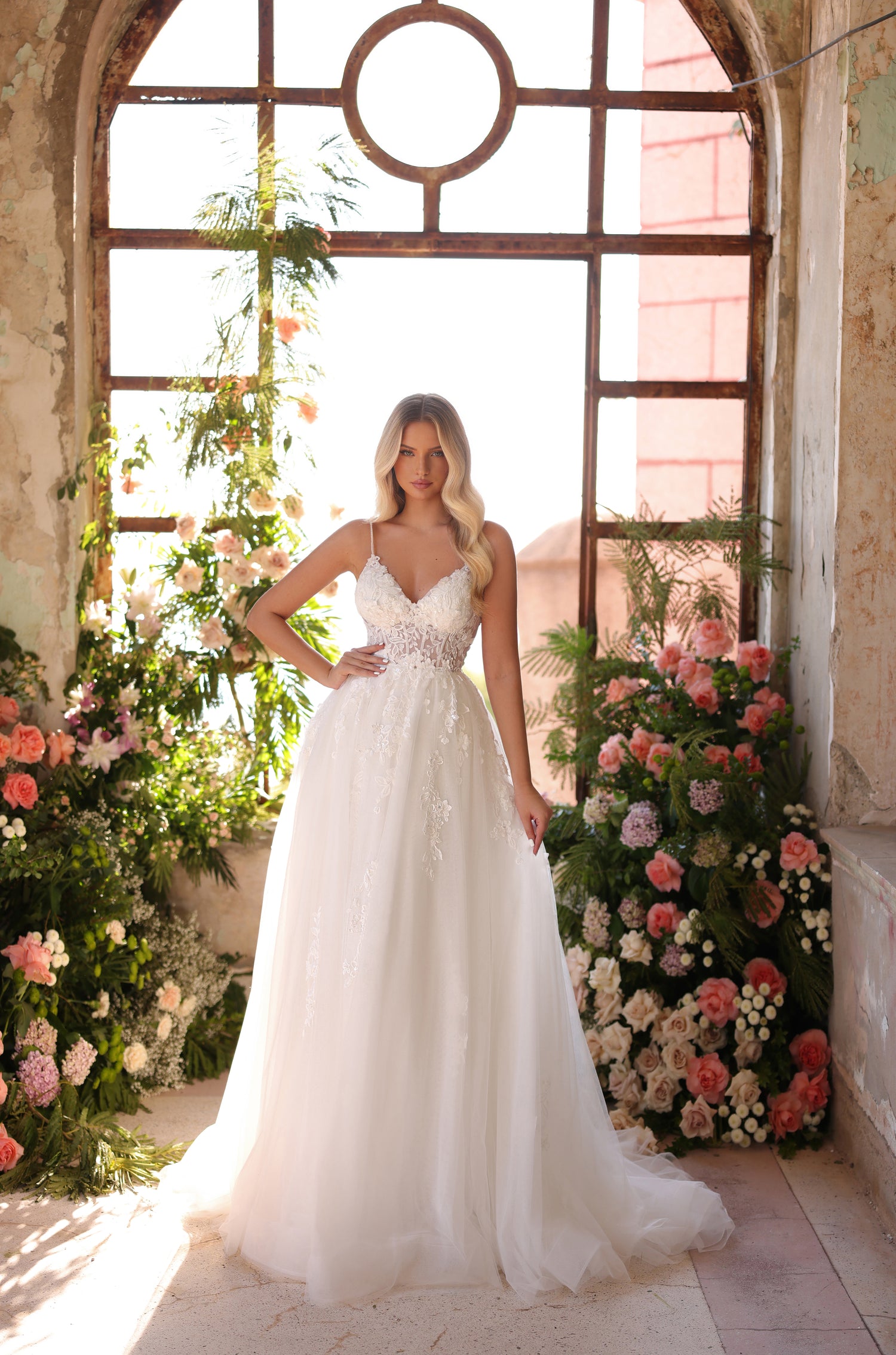 A blonde woman in a white lace wedding dress poses in a sunlit room filled with pink and white flowers.