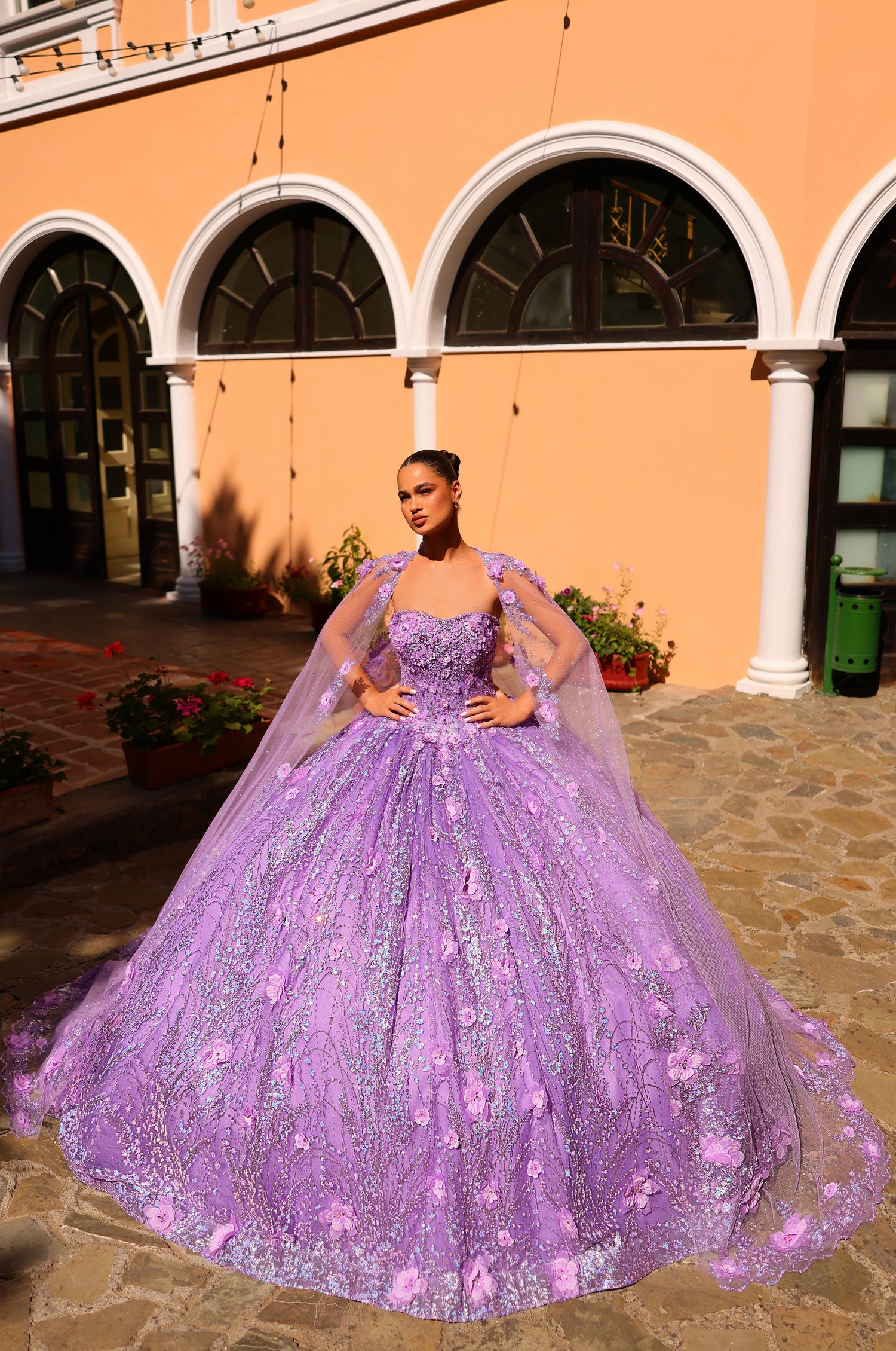 A woman wears a voluminous lilac ball gown with floral appliqués, posing in an arched courtyard.