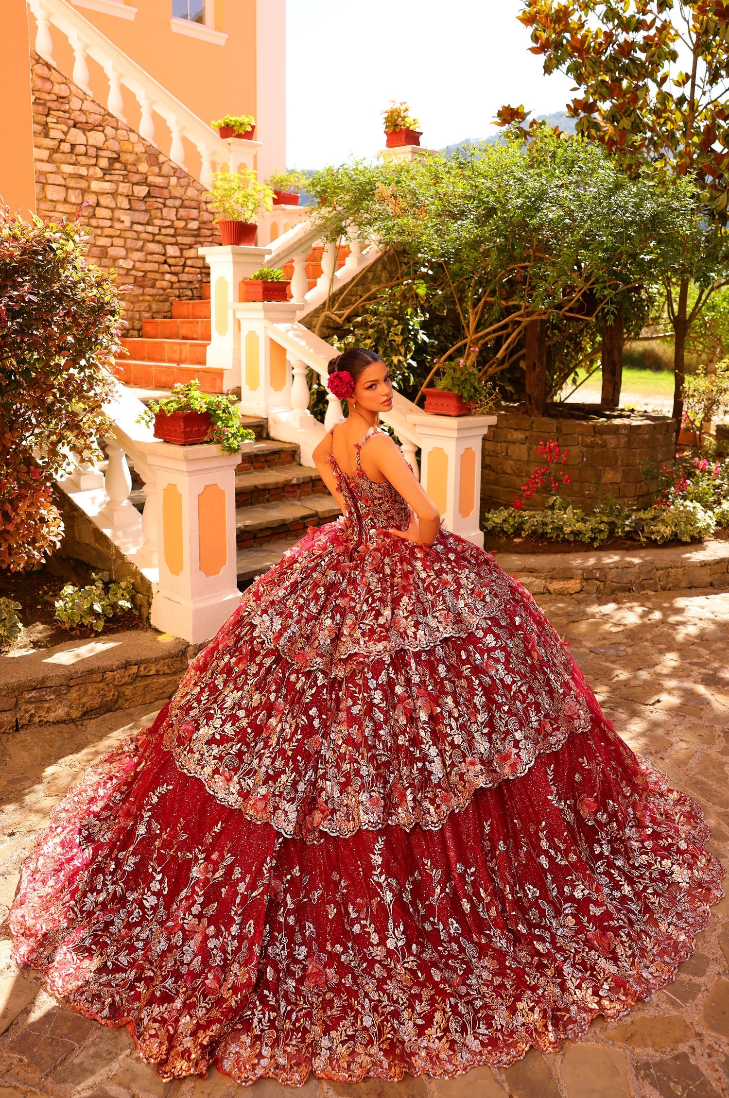 A woman in a red and silver floral quinceañera gown poses on stone steps with lush garden surroundings.