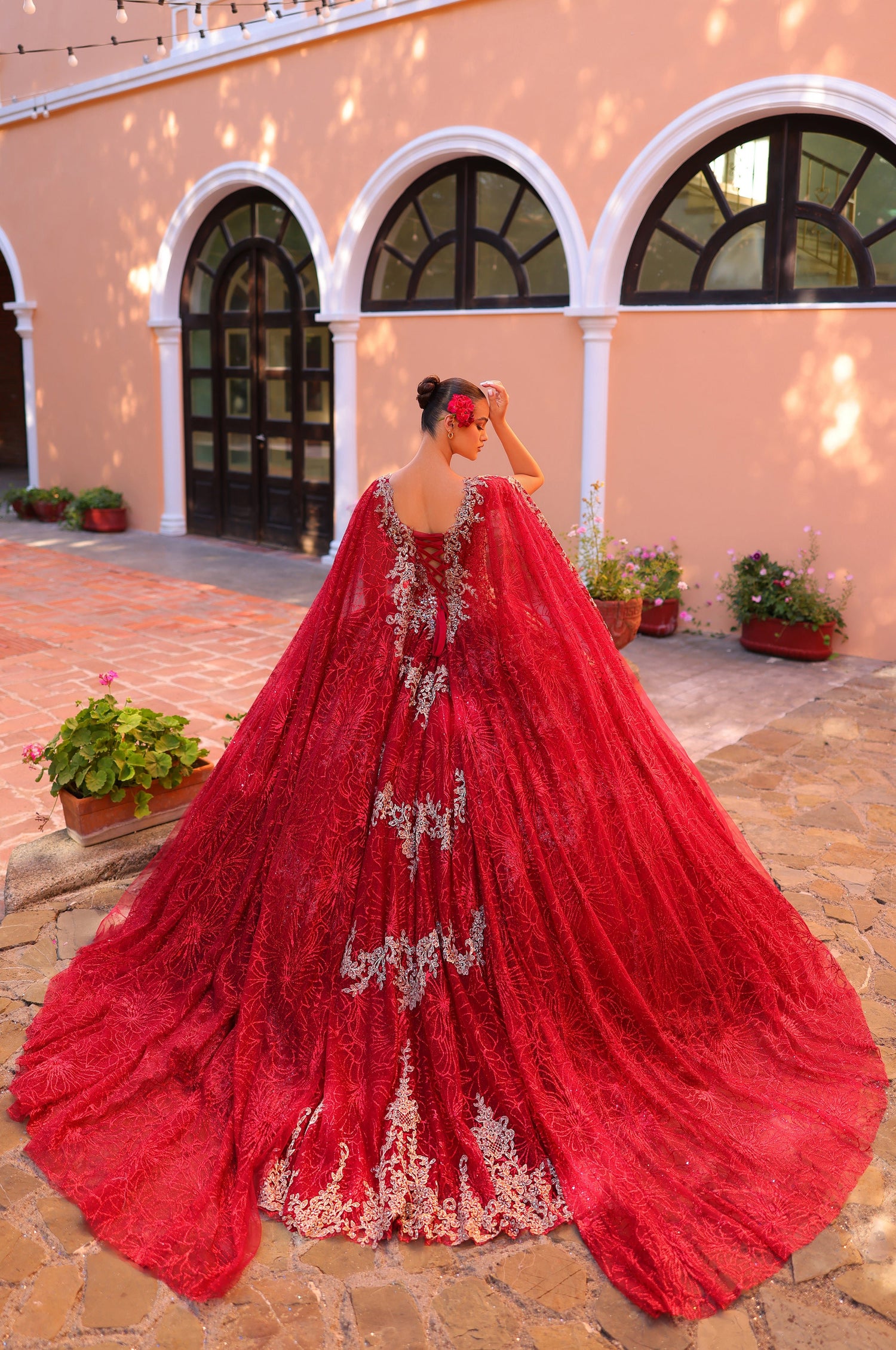 A woman in a dramatic red ball gown with silver embroidery stands in a courtyard with arched windows.