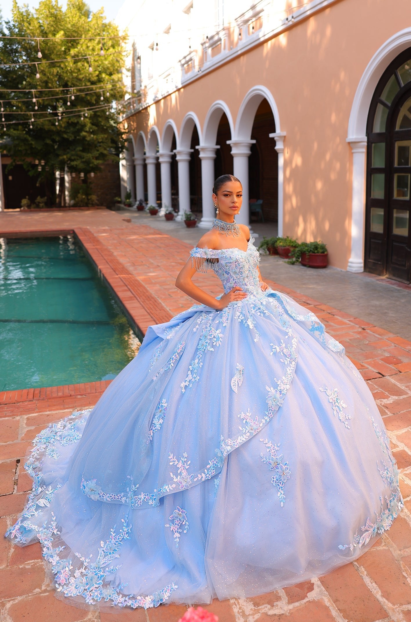A young woman wears a light blue ballgown with floral embroidery, posing by a pool near a Spanish-style building.
