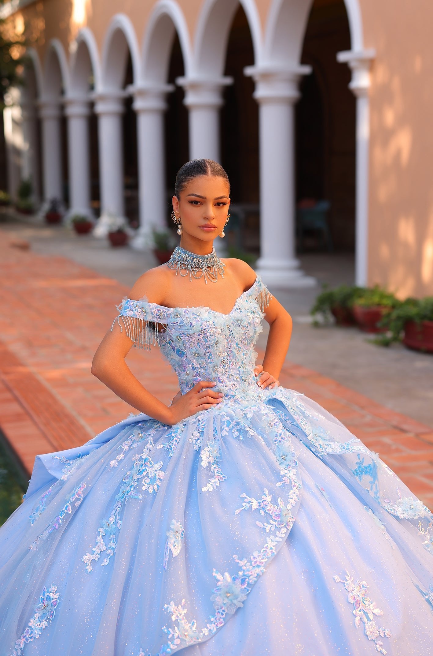 A woman in a blue ballgown with floral embellishments stands confidently in an arched columned courtyard.