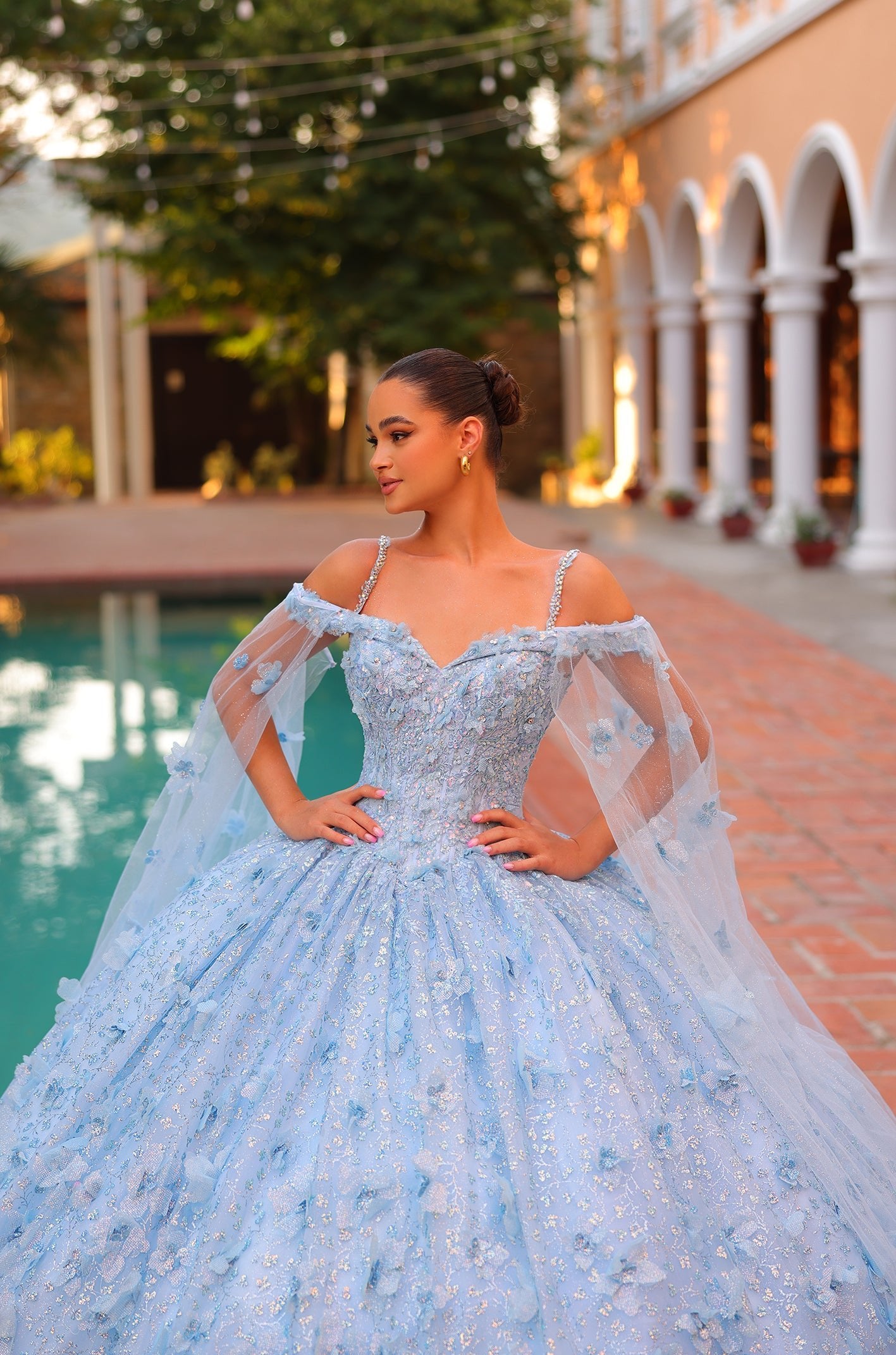 A woman in a sparkling blue ball gown stands by a pool, wearing an off-shoulder dress with delicate tulle details.