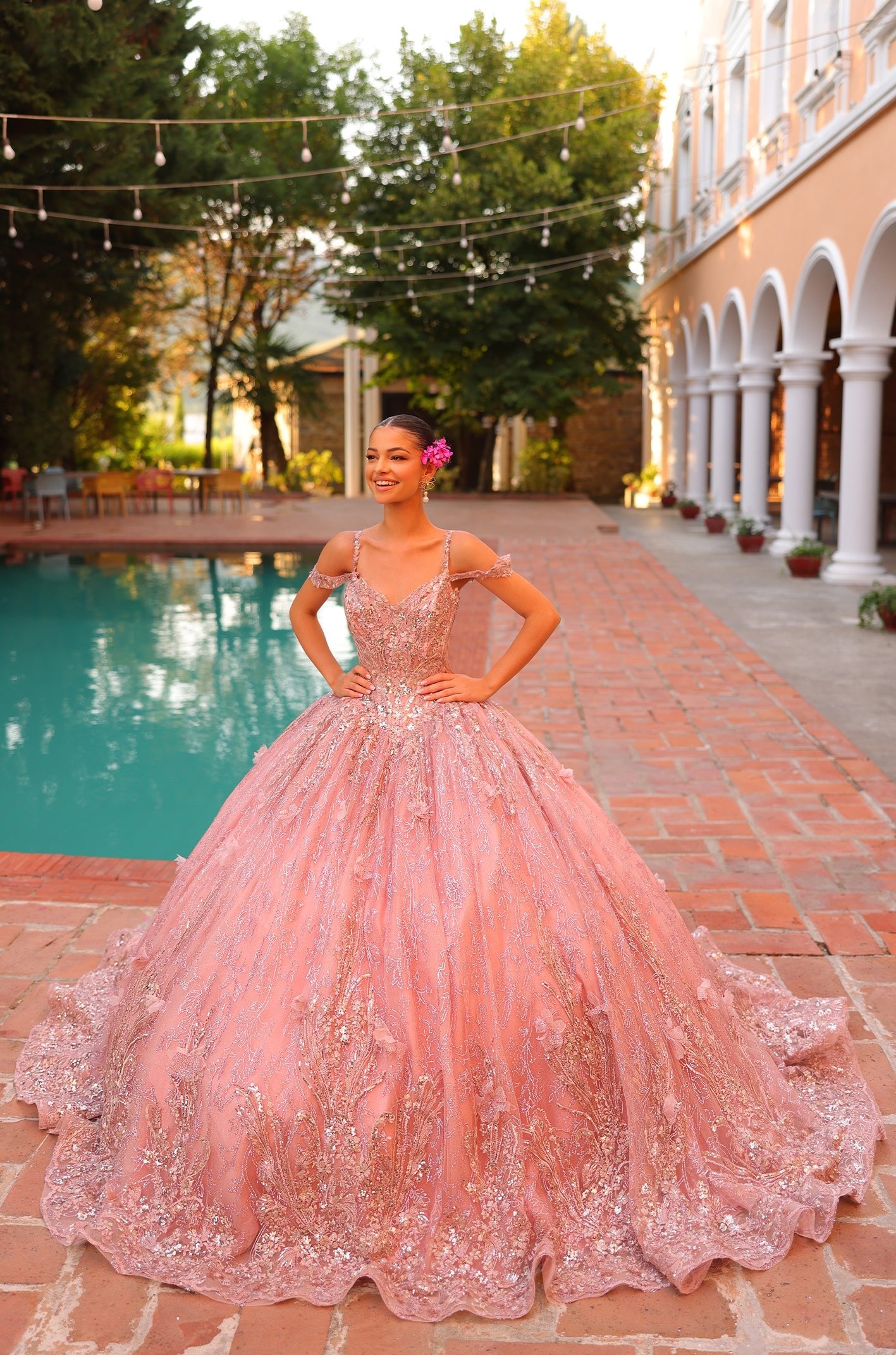 A young woman wears a blush pink, glittery ball gown by a sparkling pool with string lights overhead.