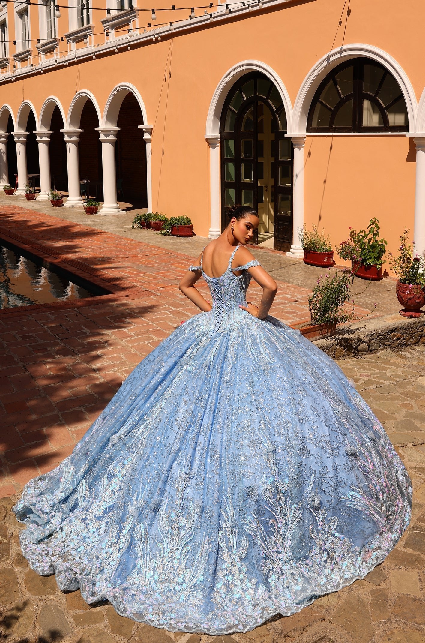 A blue ballgown with intricate lace stands against an arched, terracotta-colored building with stone walkway.