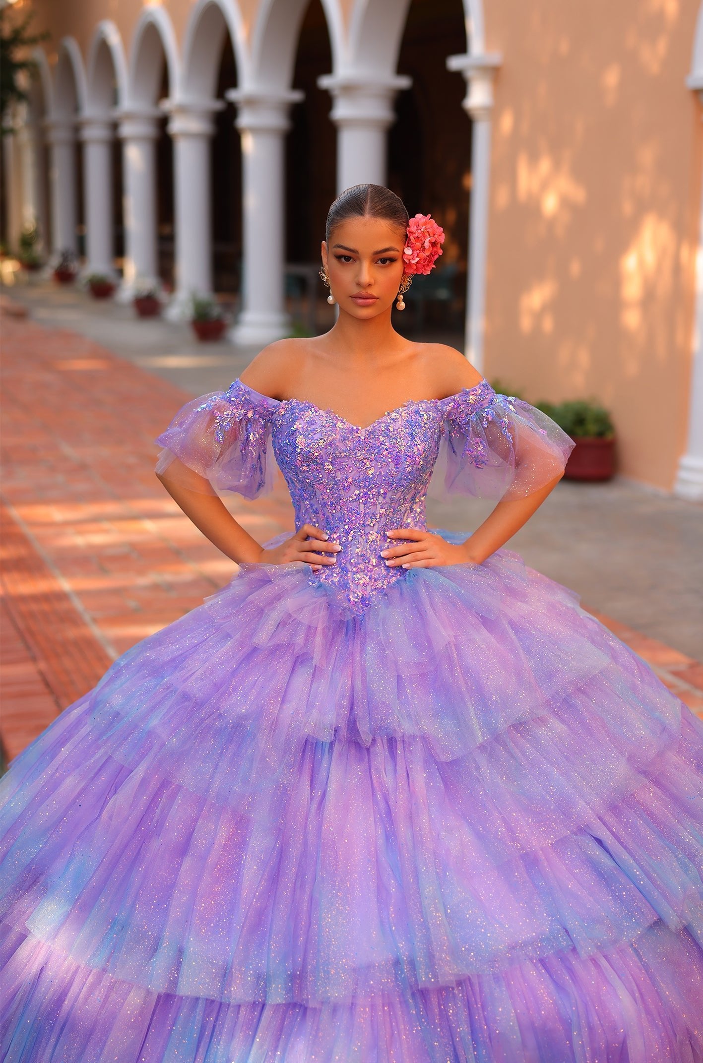 A woman in a sparkling lavender ball gown poses confidently with a coral flower in her hair.