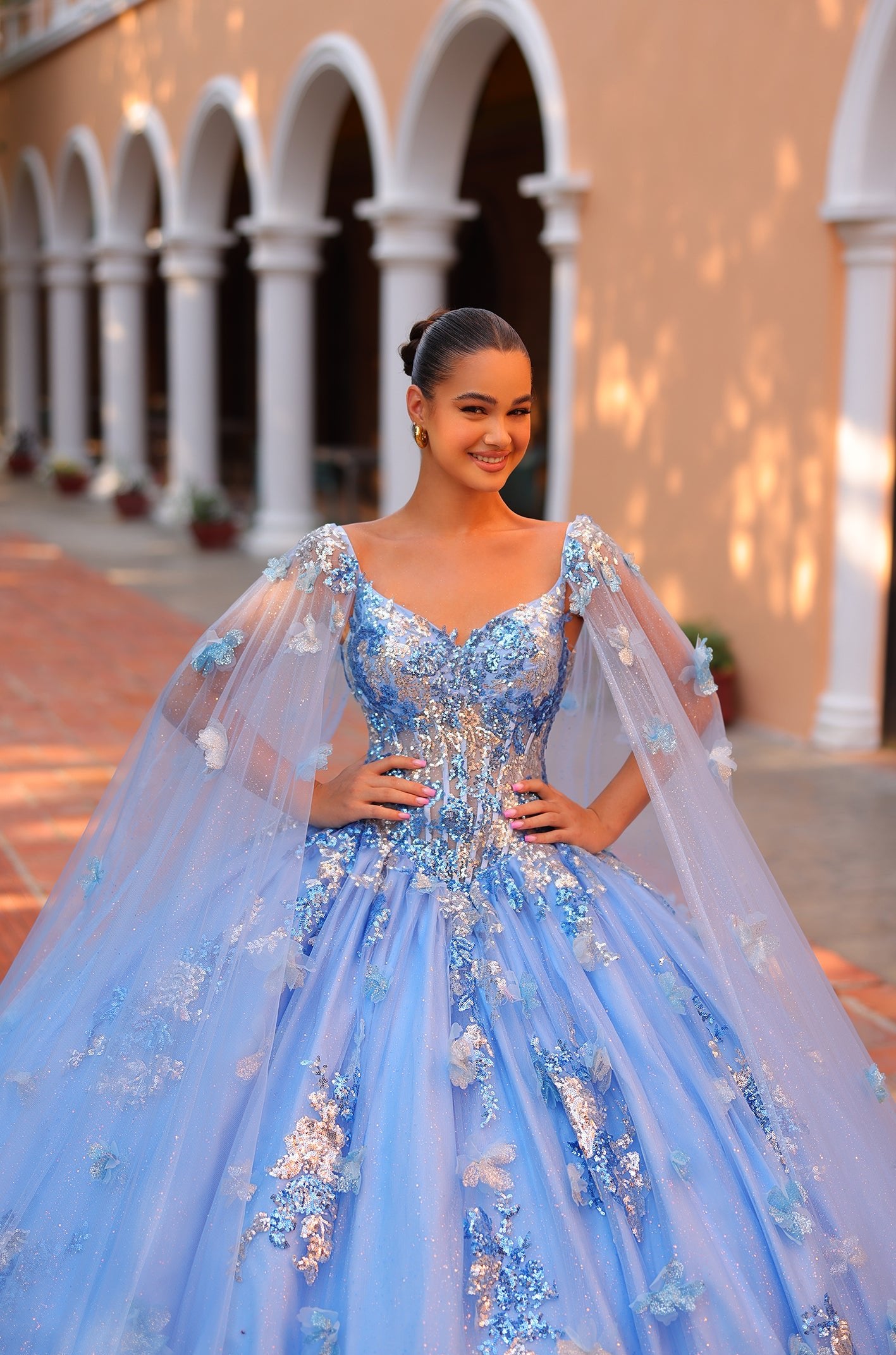A young woman wears a blue ballgown with shimmering floral details, standing confidently in an arched architectural setting.