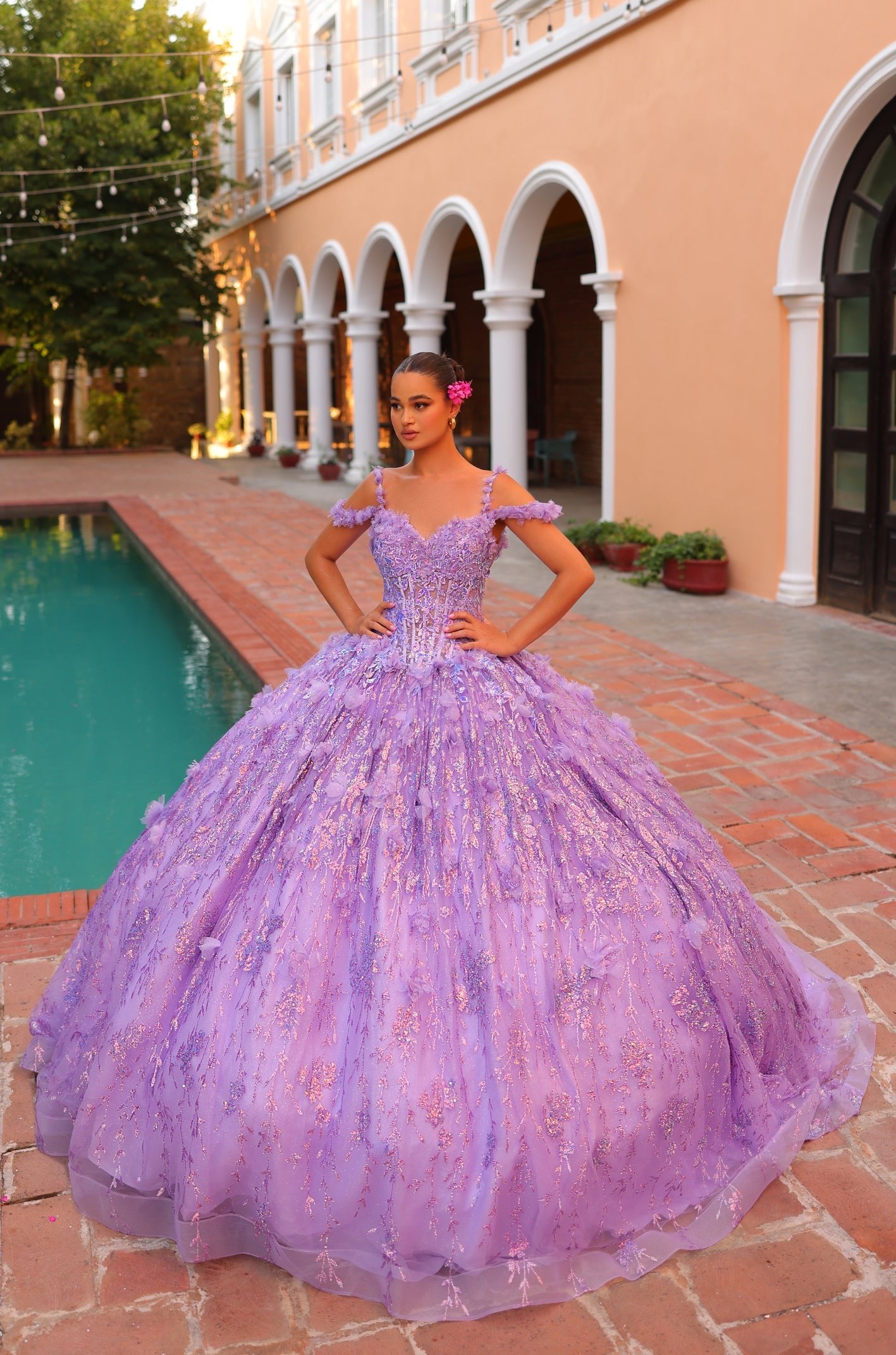 A model wears a voluminous lilac quinceañera ball gown with sparkly details near a pool and arched building.