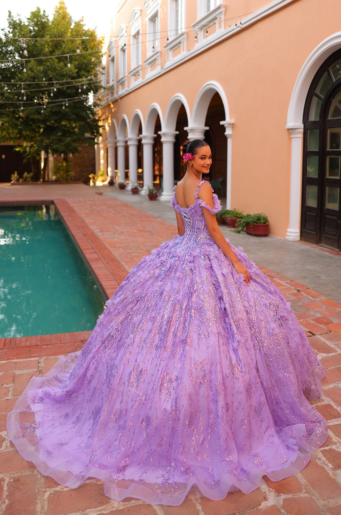 A lavender ball gown with floral details on a woman standing by a pool near an arched building.