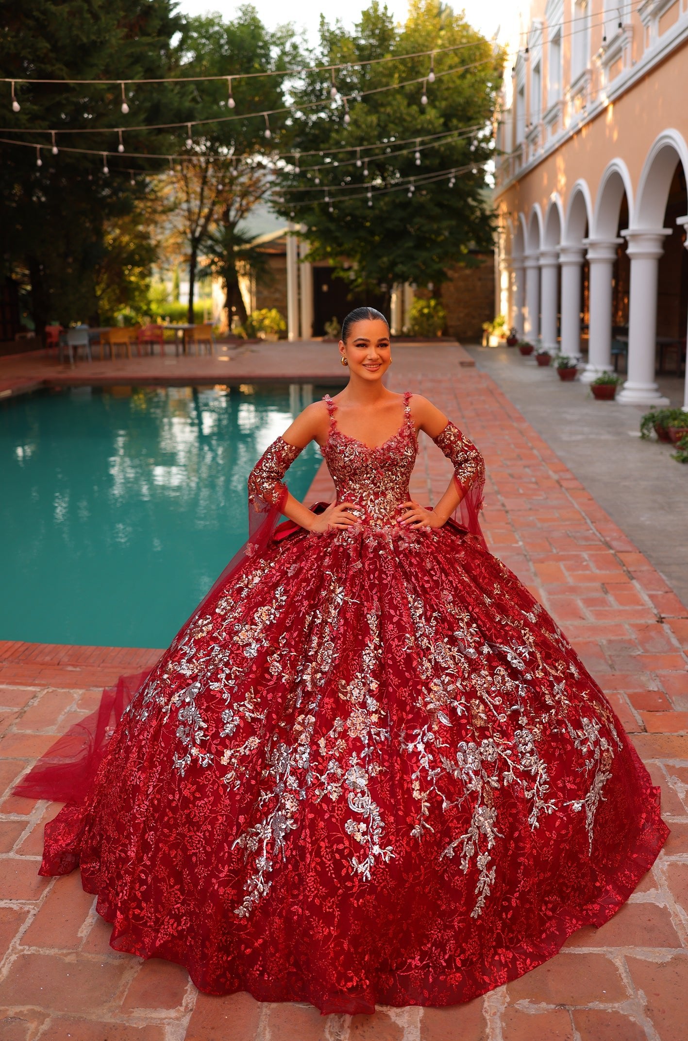 A Red/Multi ball gown with silver embellishments stands beside a pool, worn by a smiling person in a scenic setting.
