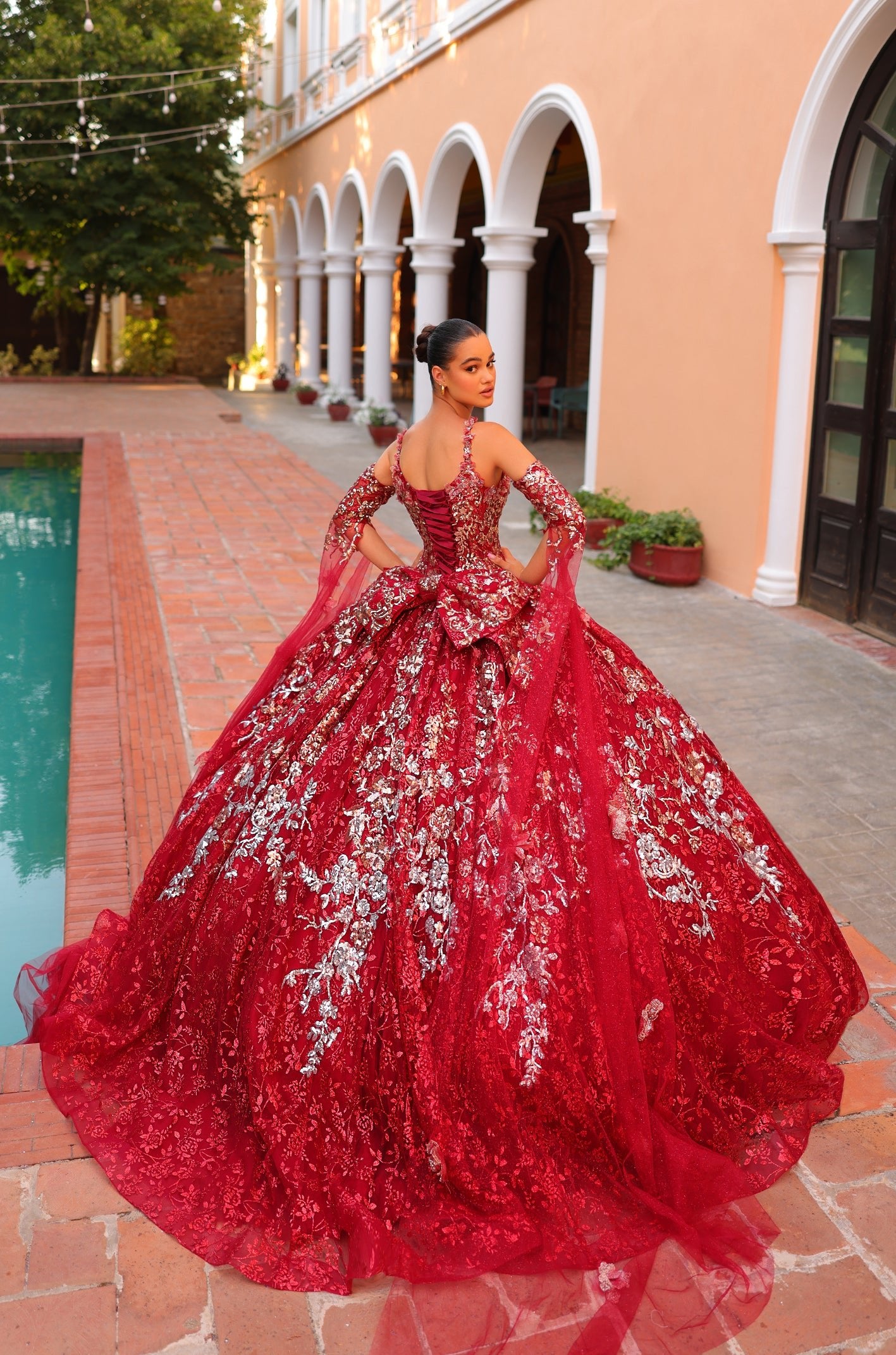 A woman wears an elaborate red and silver quinceañera ball gown with floral embroidery near a poolside venue.