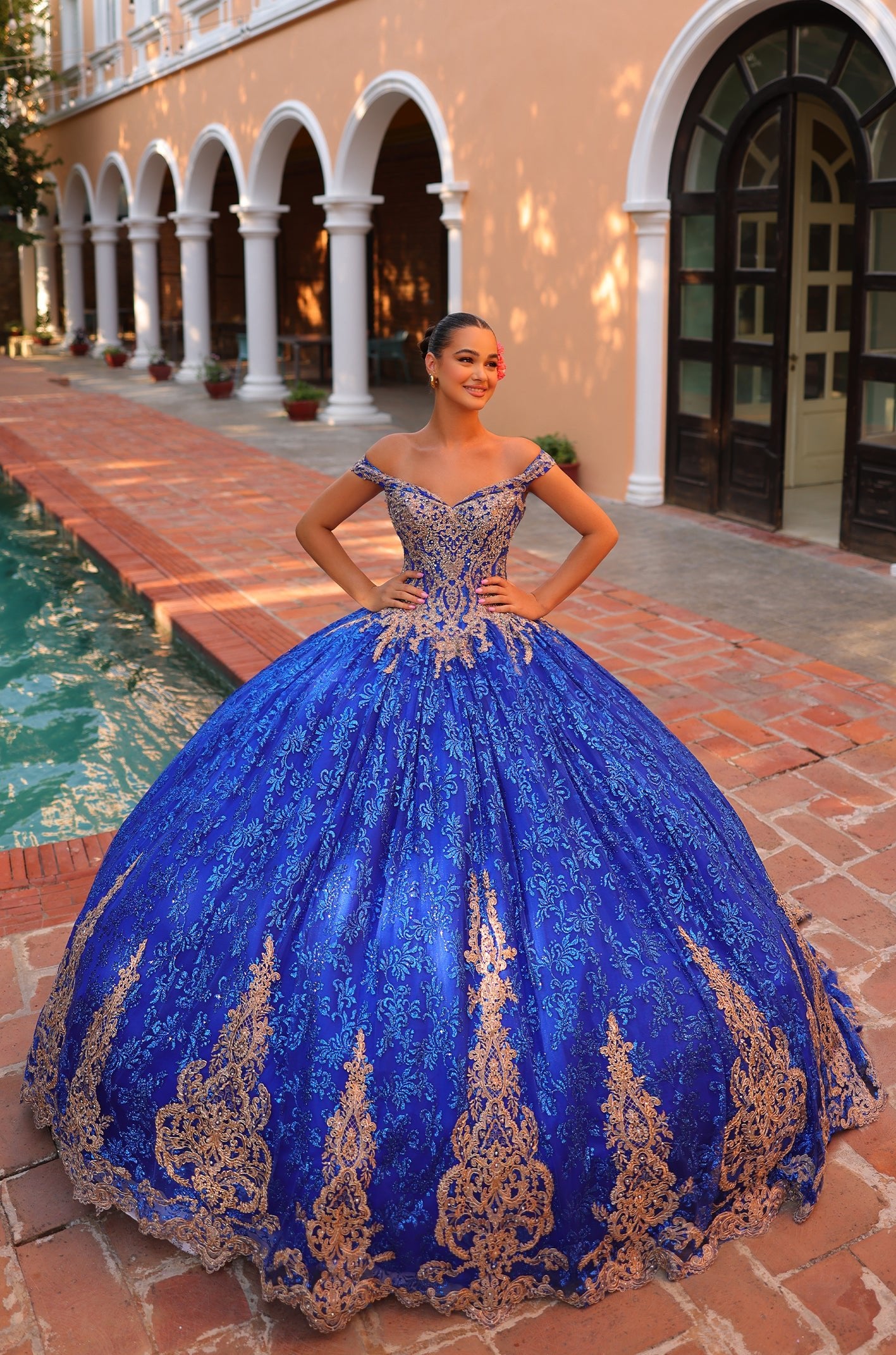 A young woman wears an elaborate Royal Blue/Gold quinceañera ball gown with intricate lace detailing in an arched courtyard.