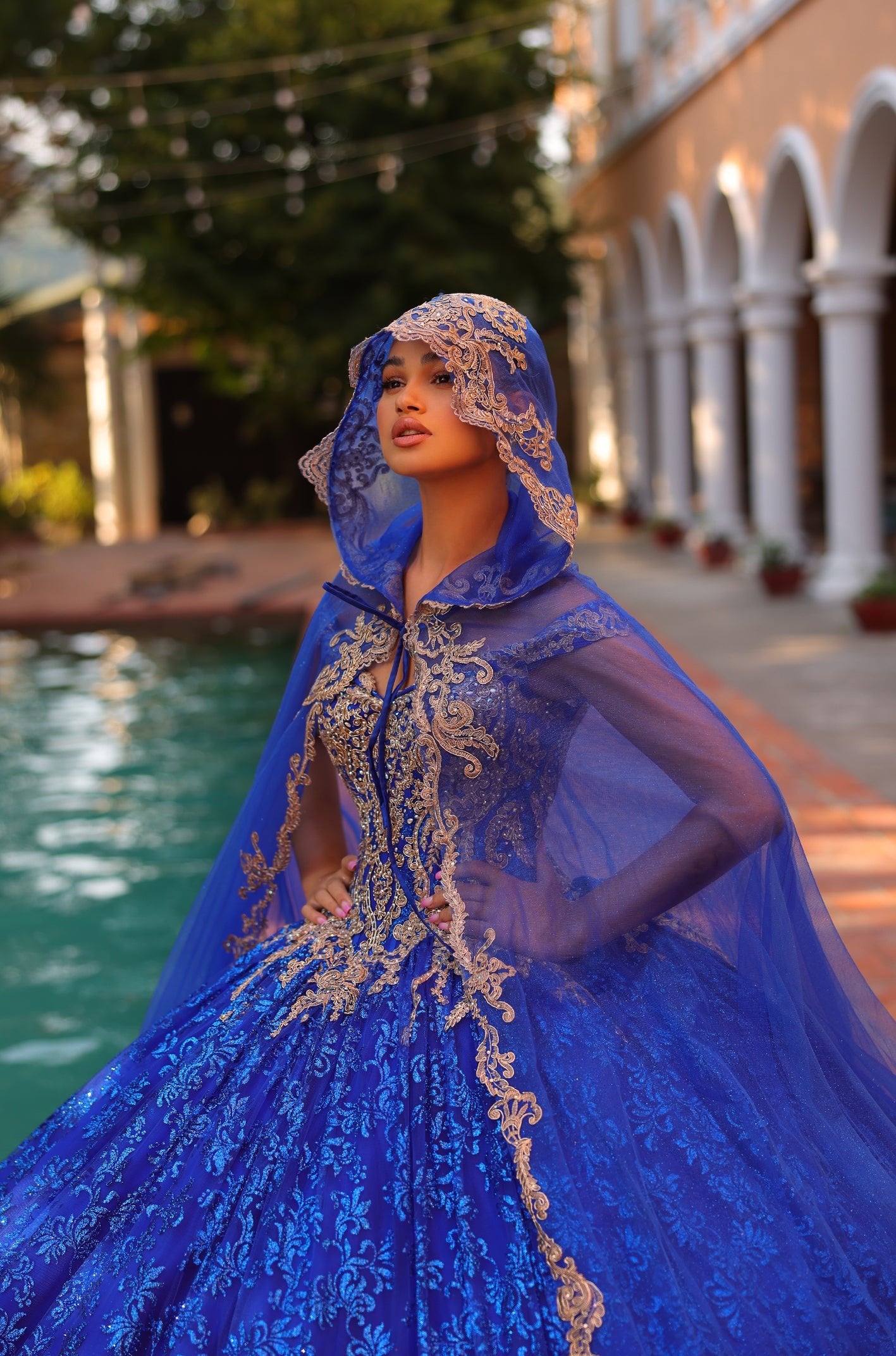 A woman in an elaborate blue ball gown with gold embroidery stands by a pool, wearing a matching hooded cape.